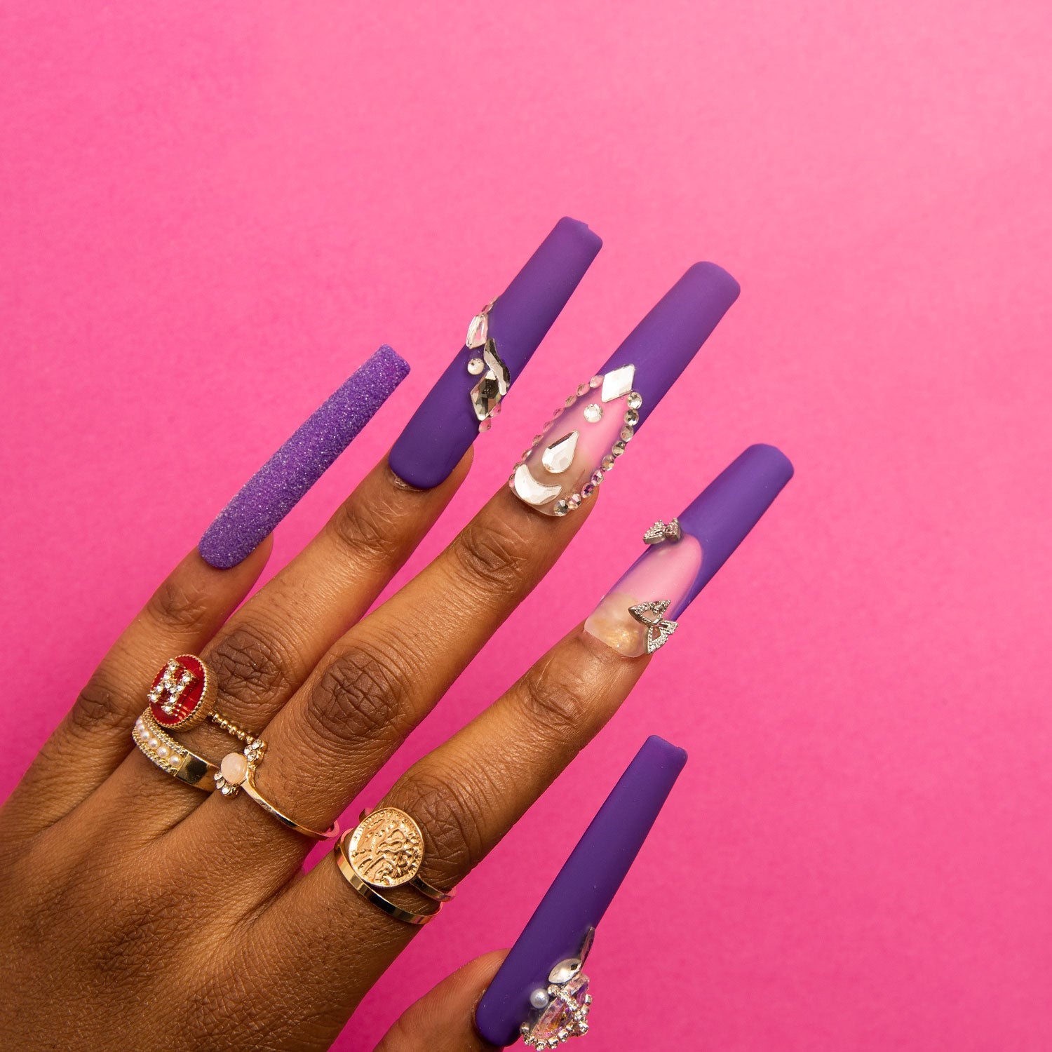 Hand showcasing 'Mystique Veil' press-on nails by Lovful, featuring deep purple base with butterfly decorations, gem accents, and glitter overlay against a pink background.