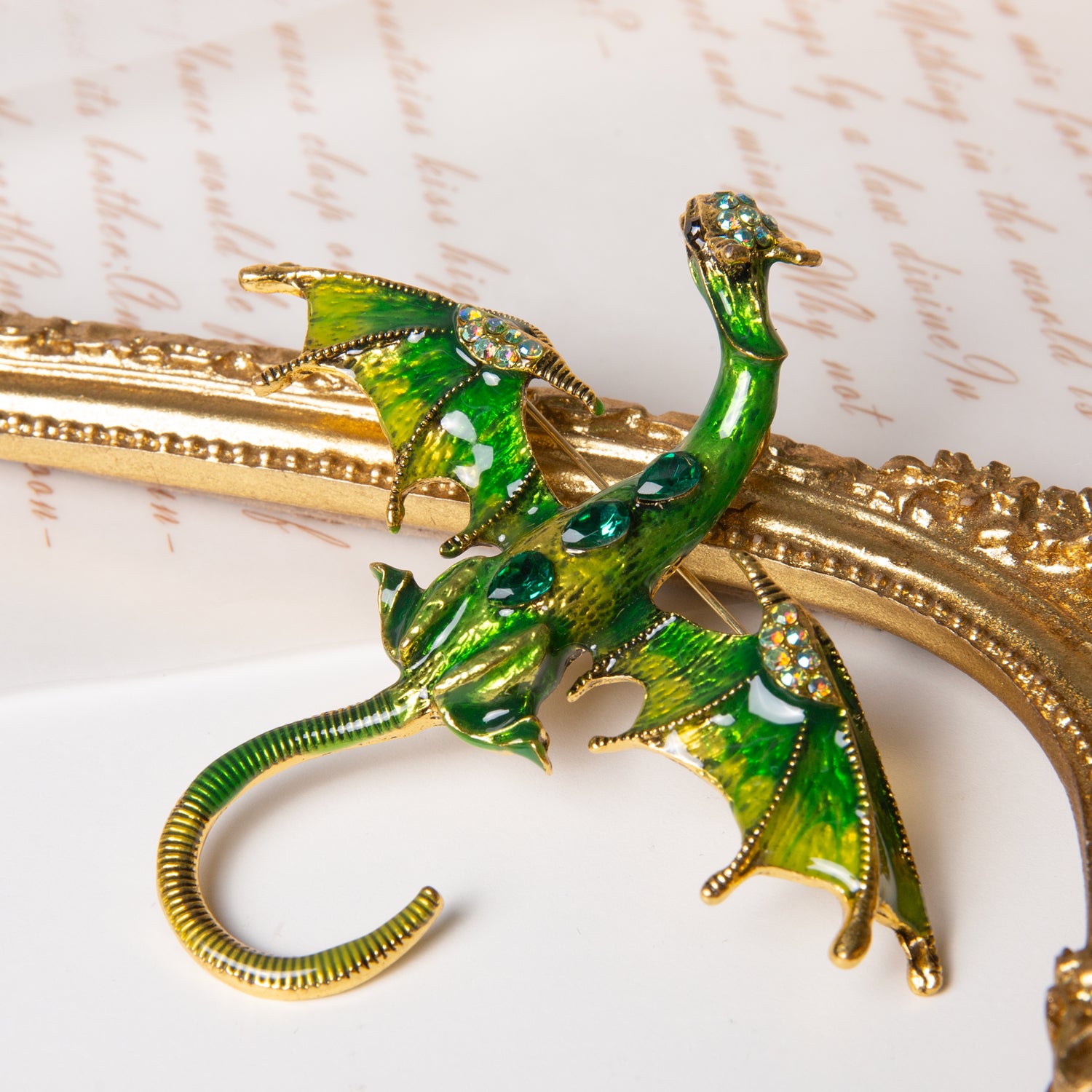 Close-up of Enchanting Fairy Queen dragon brooch with green and gold details, displayed on ornate gold frame and cursive script background.