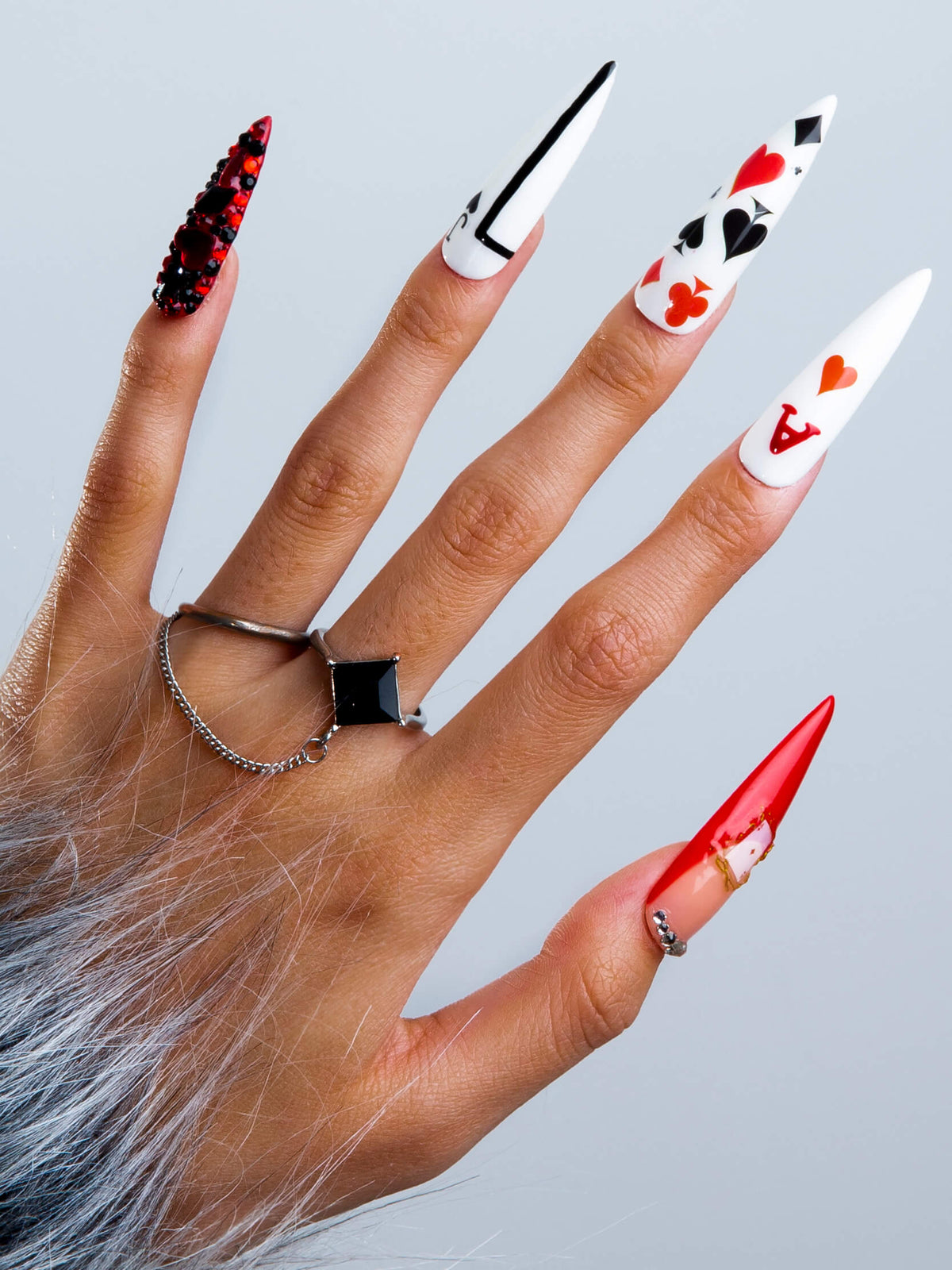 Hand with Poker Queen stiletto-shaped press-on nails featuring red and white playing card designs. Nails decorated with hearts and card suits, worn with silver rings.