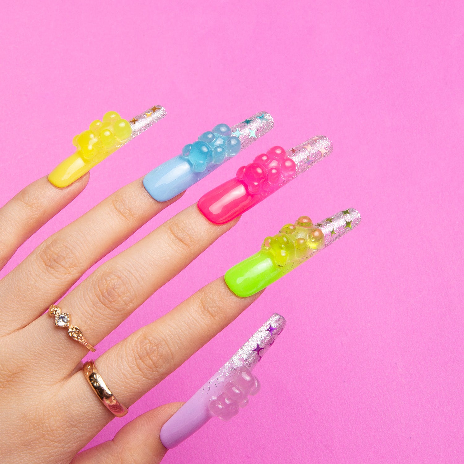 Hand wearing 'Sweet Wonderland' press-on nails by Lovful.com. Nails in yellow, blue, pink, green, and purple with glittery tips and gummy bear charms against a pink background.