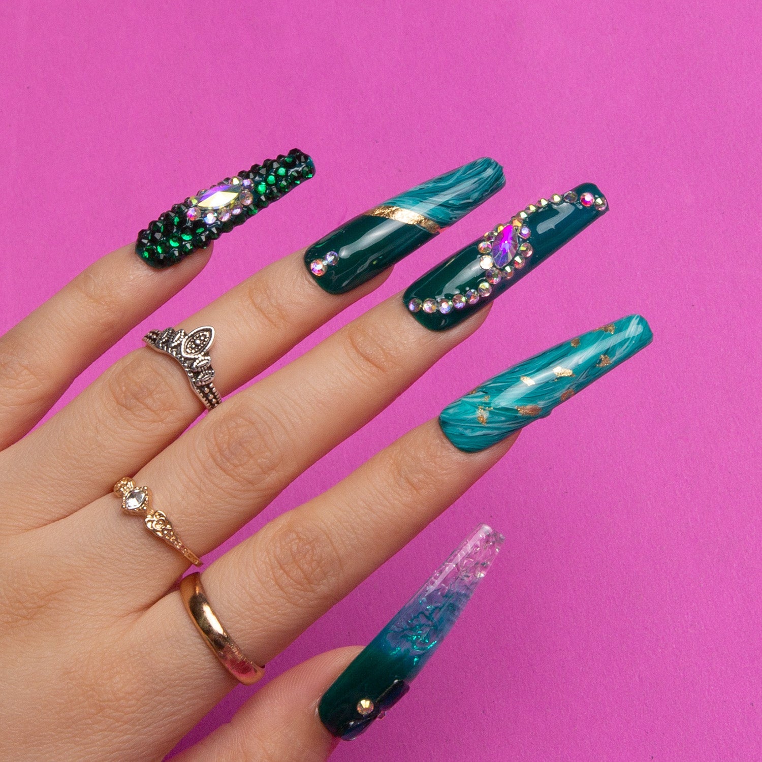 Hand showcasing Lovful's 'Emerald Envy' Coffin-shaped press-on nails with green designs, gold decor, and green rhinestones against a pink background.