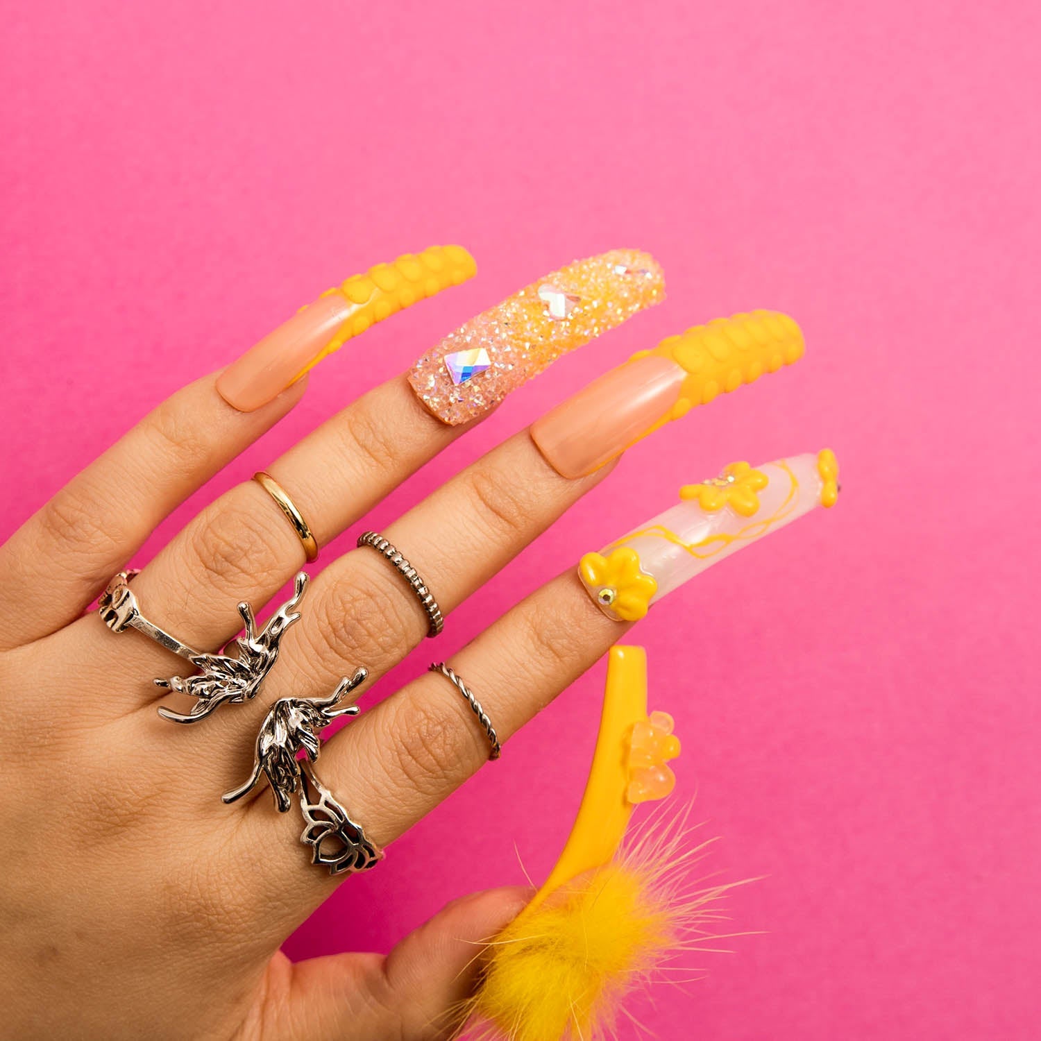 Hand with Lovful.com's H131 - Yummy - Curve - RTS press-on acrylic nails in sunny yellow, decorated with glitter and detachable fluffy balls, set against a bright pink background, with several rings on fingers.
