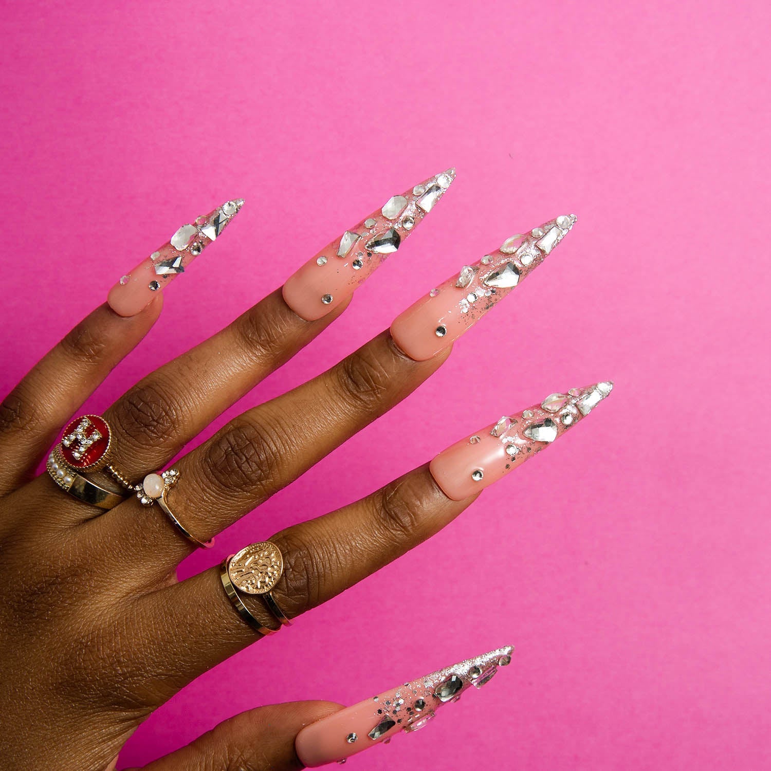 Hand displaying stiletto-shaped 'Silver Wind' press-on nails by Lovful, with sparkling silver glitter, rhinestones, and multiple rings, against a pink background