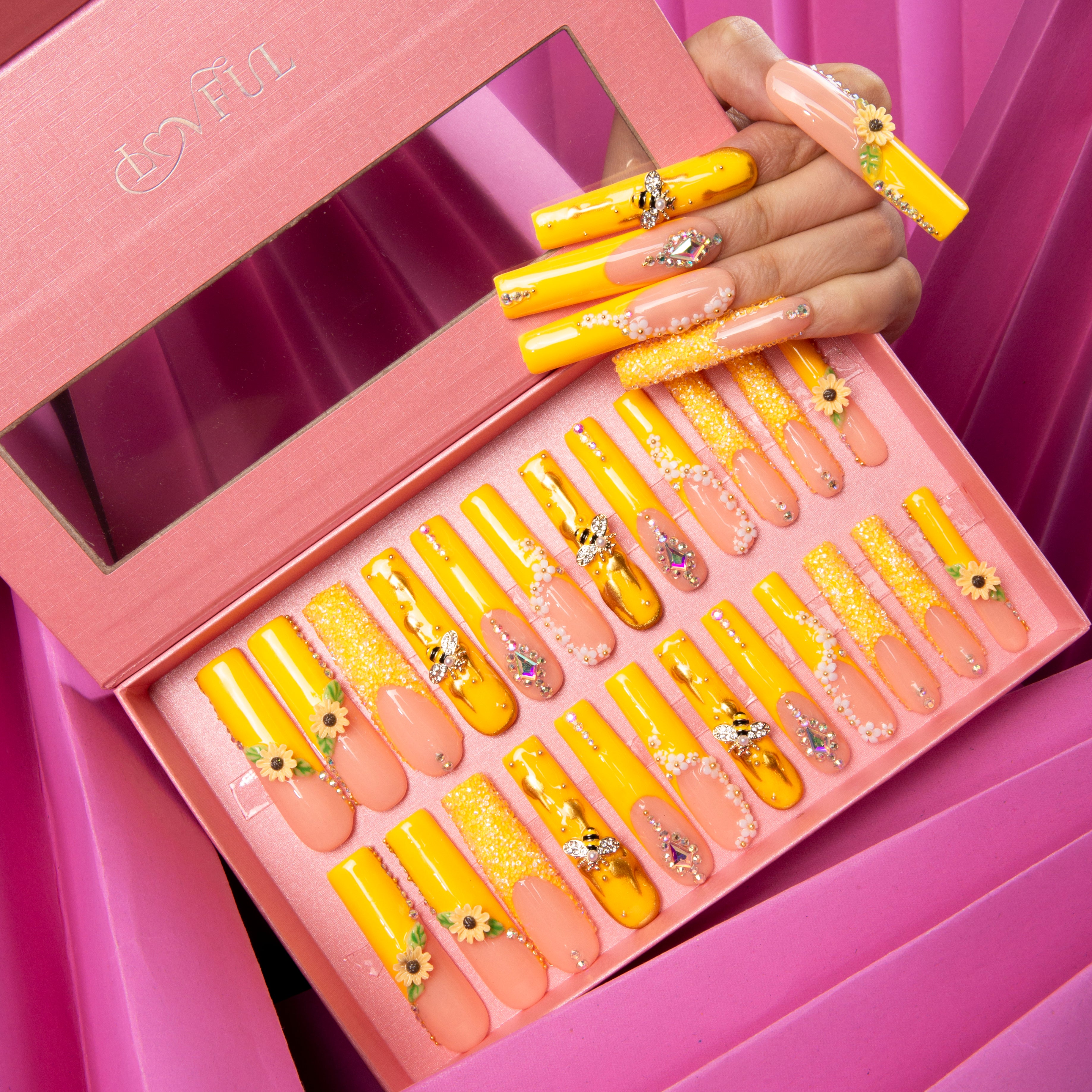 Box of 24 press-on acrylic nails with bright yellow and pink gradient, sunflowers and bee accents. A hand displaying the nails shows the detailed design.