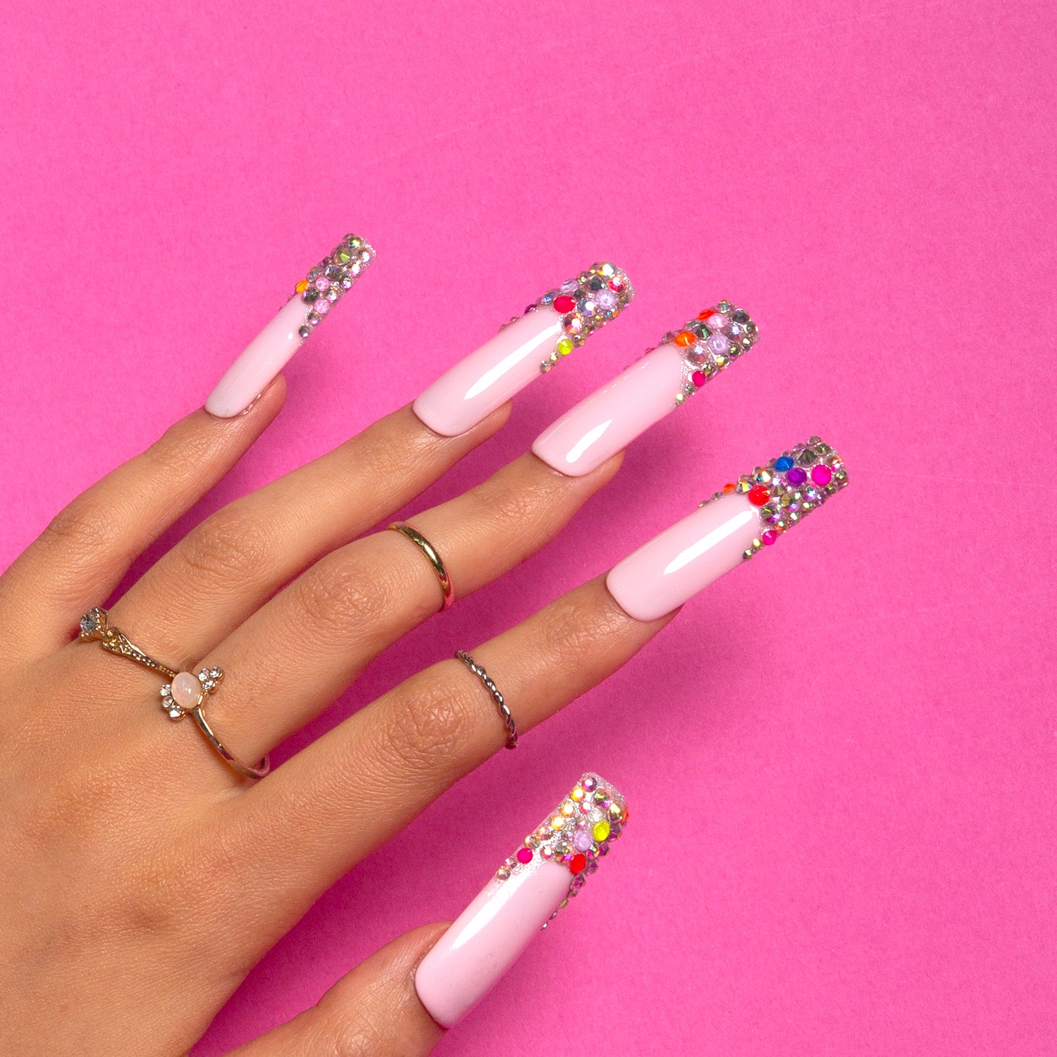 Hand showcasing Lovful 'Jelly Bean' Press-On Nails with nude base and colorful beans on the tips, set against a pink background. Hand adorned with gold rings for added elegance.