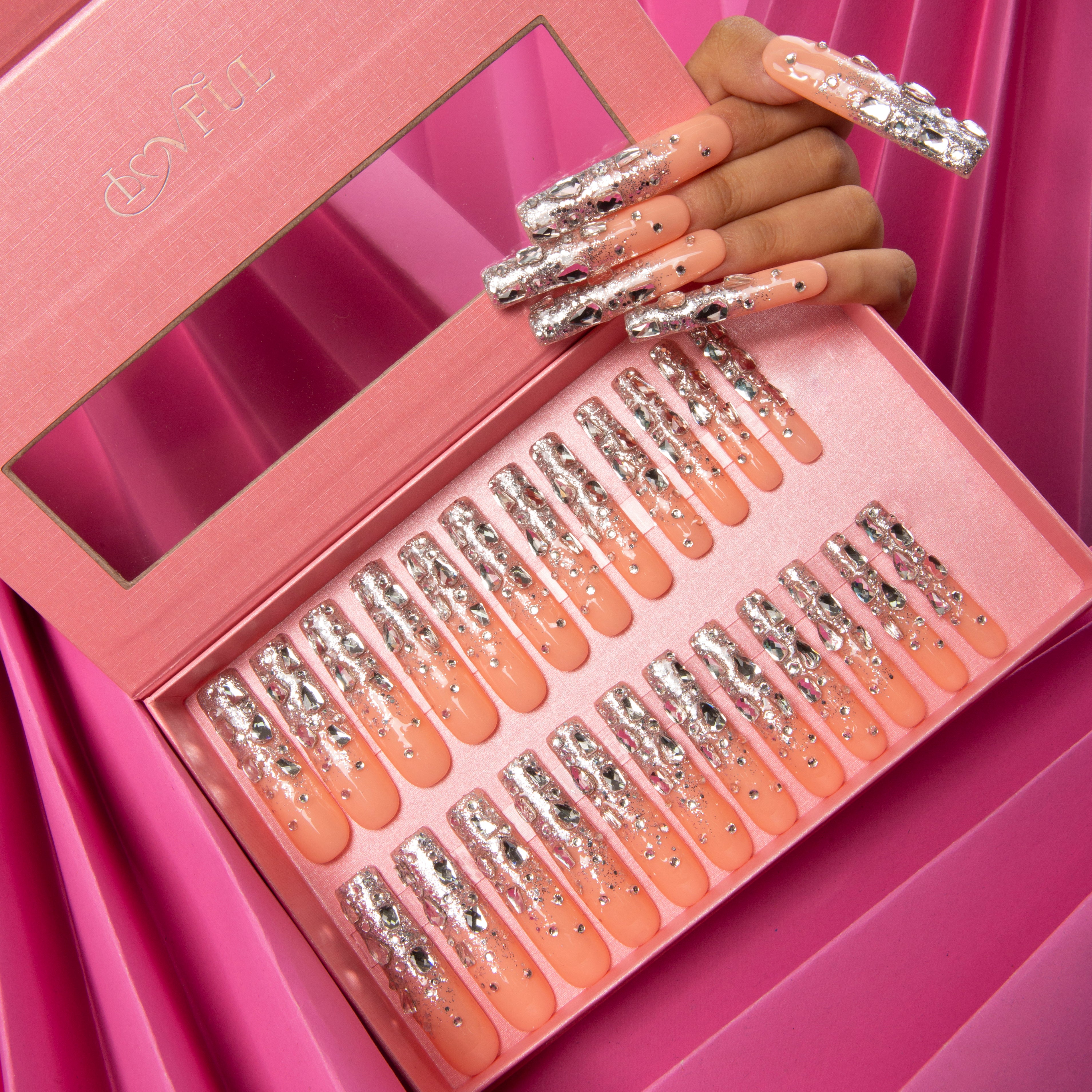 'Silver Wind' press-on acrylic nails - 24 pieces in a pink box, featuring nude base with silver glitter and rhinestones. Hand displaying applied nails on a pink background.