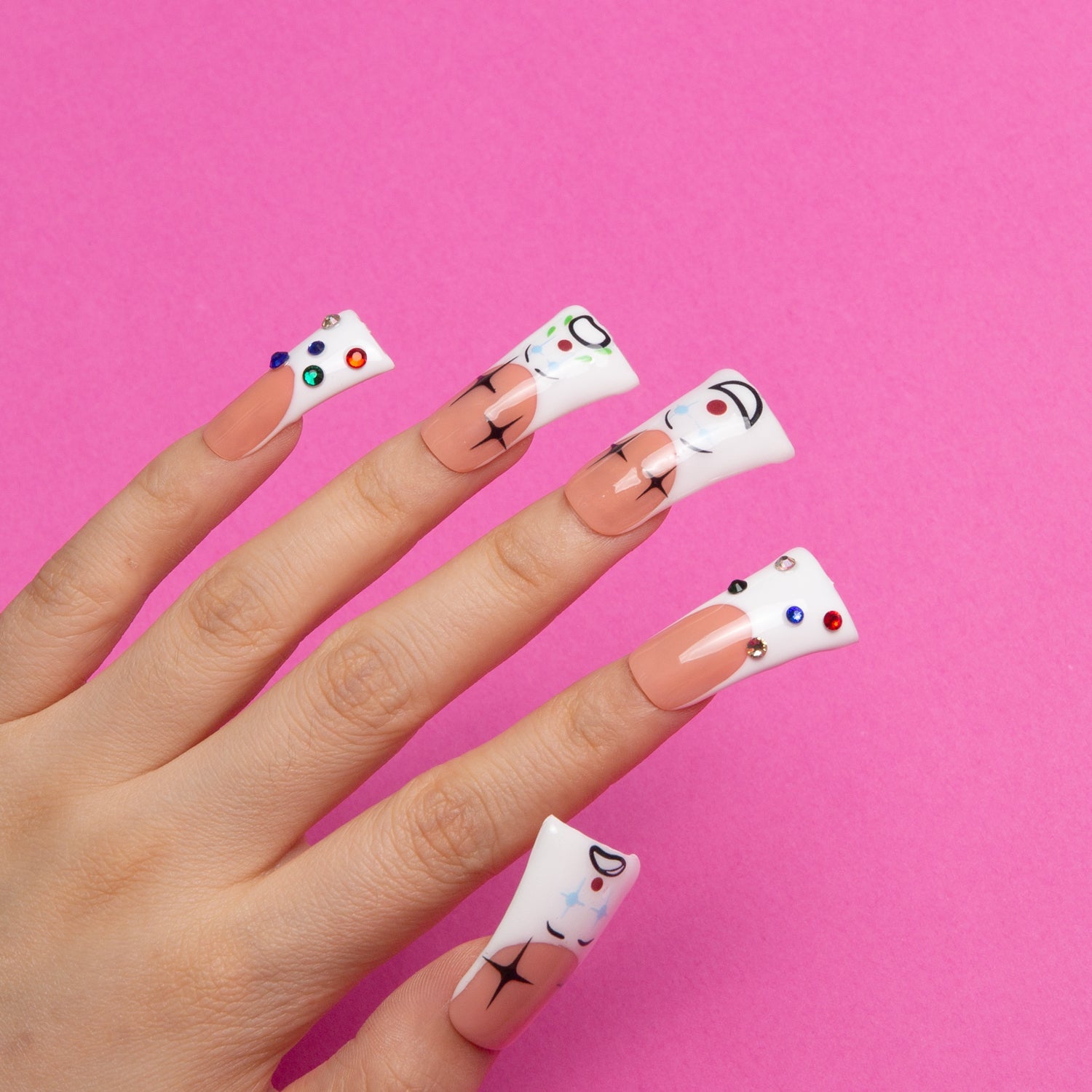 Hand with circus clown-themed press-on nails featuring white tips, colorful rhinestones, black crossed lines, and cartoon faces on a pink background from Lovful.com.