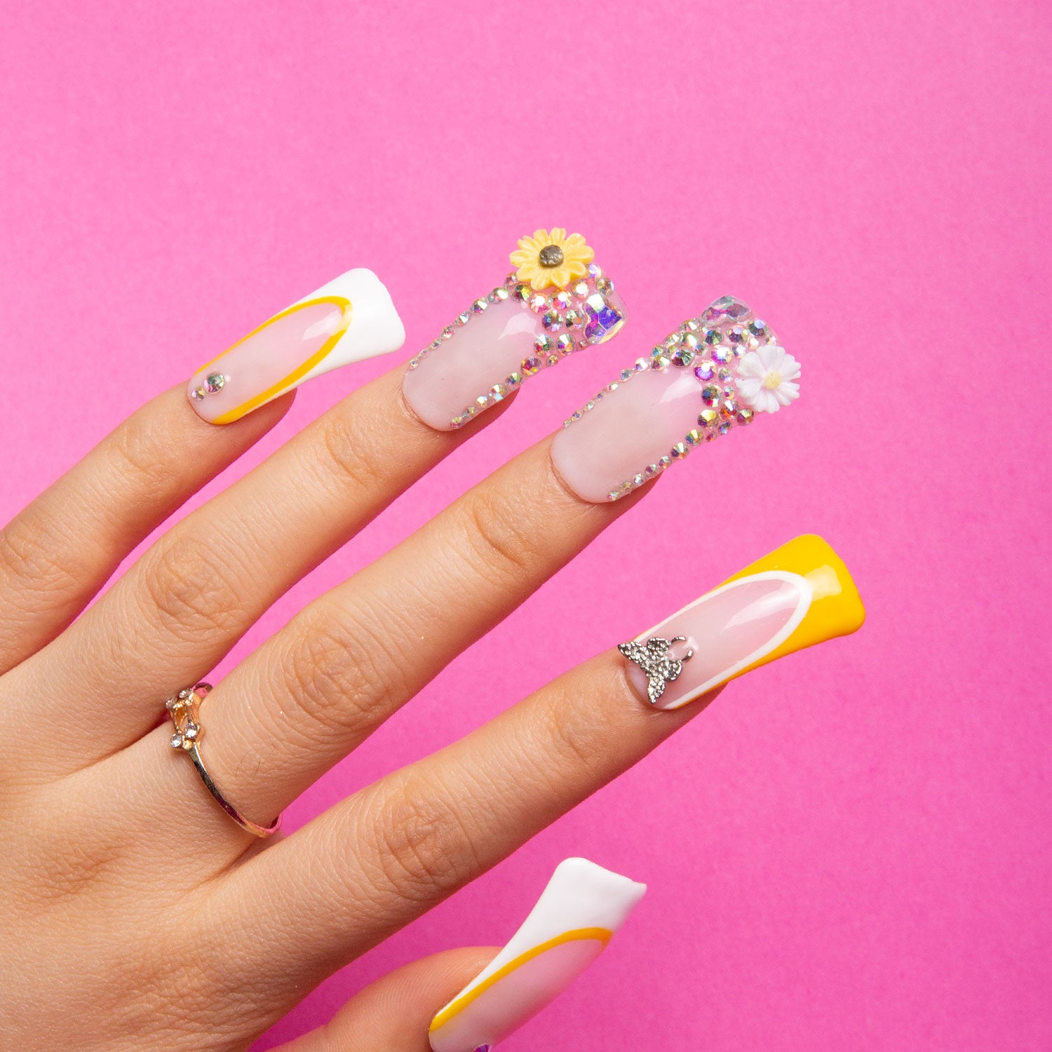 Hand with 'Sunflower' press-on nails featuring yellow hues, white French tips, sunflowers, and rhinestones over pink background