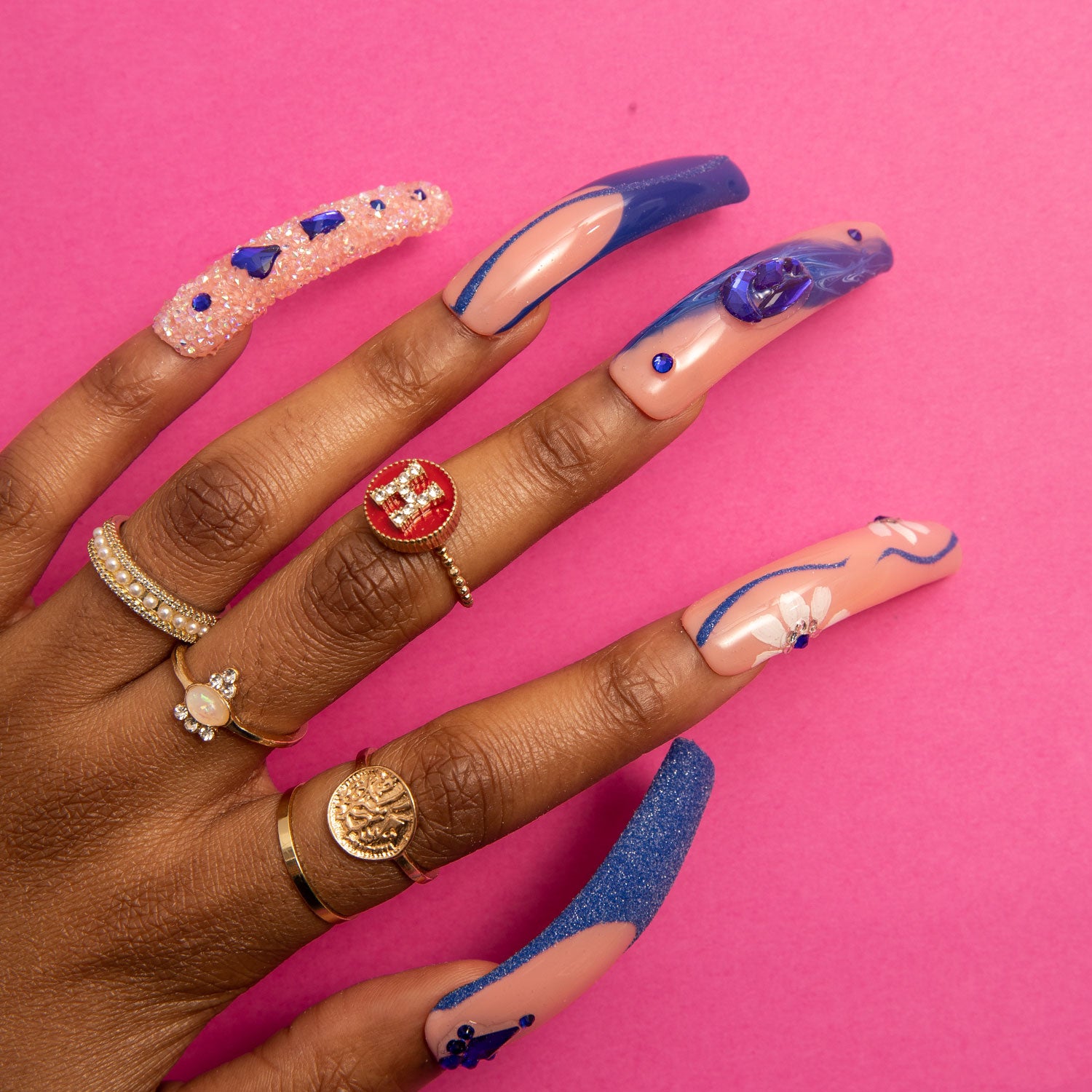 Close-up of hand with 'Blue Suede' press-on nails featuring blue French tips, curvy line designs, crystal-like decorations, and blue heart-shaped gems; hand is adorned with various stylish rings, against a pink background.