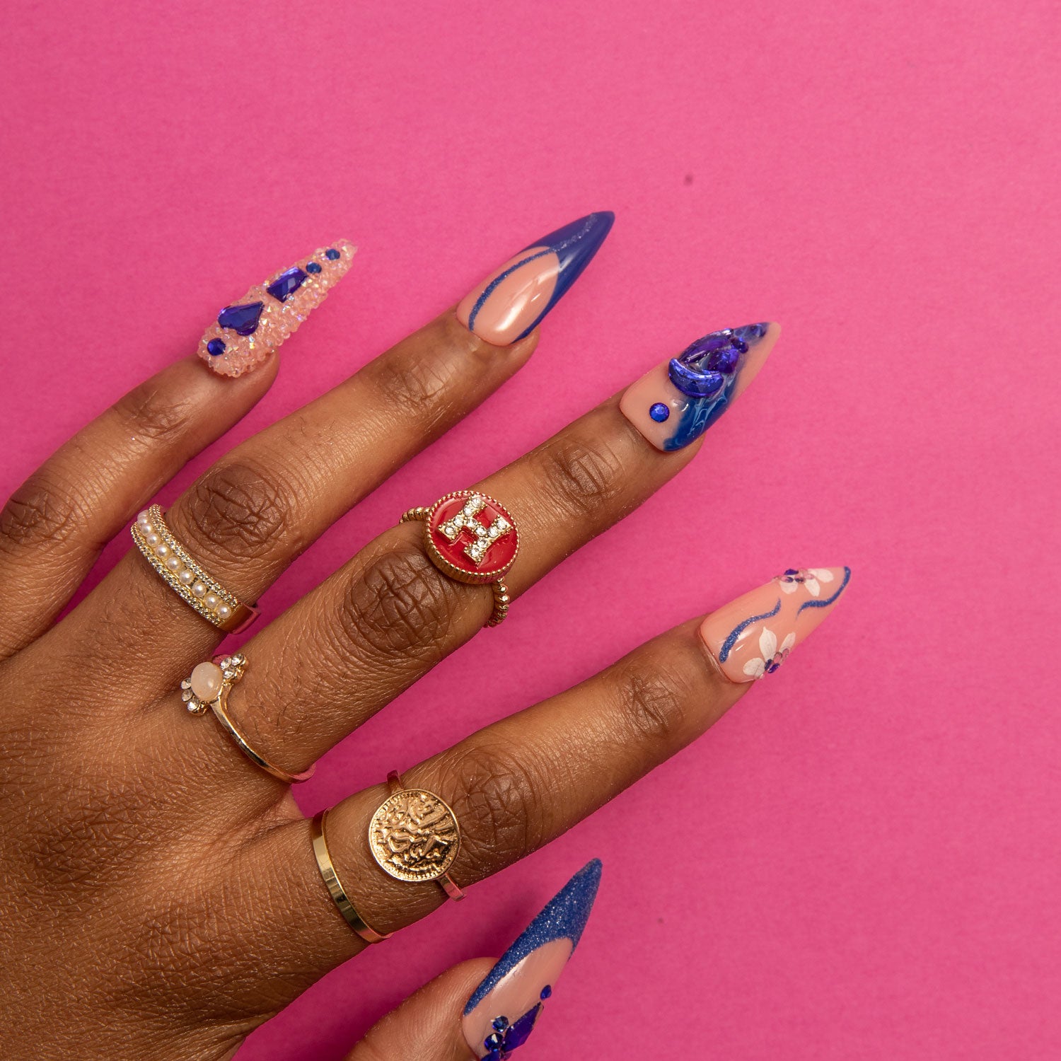 Hand with 'Blue Suede' press-on stiletto nails featuring deep blue French tips, curvy line designs, blue heart-shaped gems, crystal decorations, and adorned with gold and diamond rings against a pink background.
