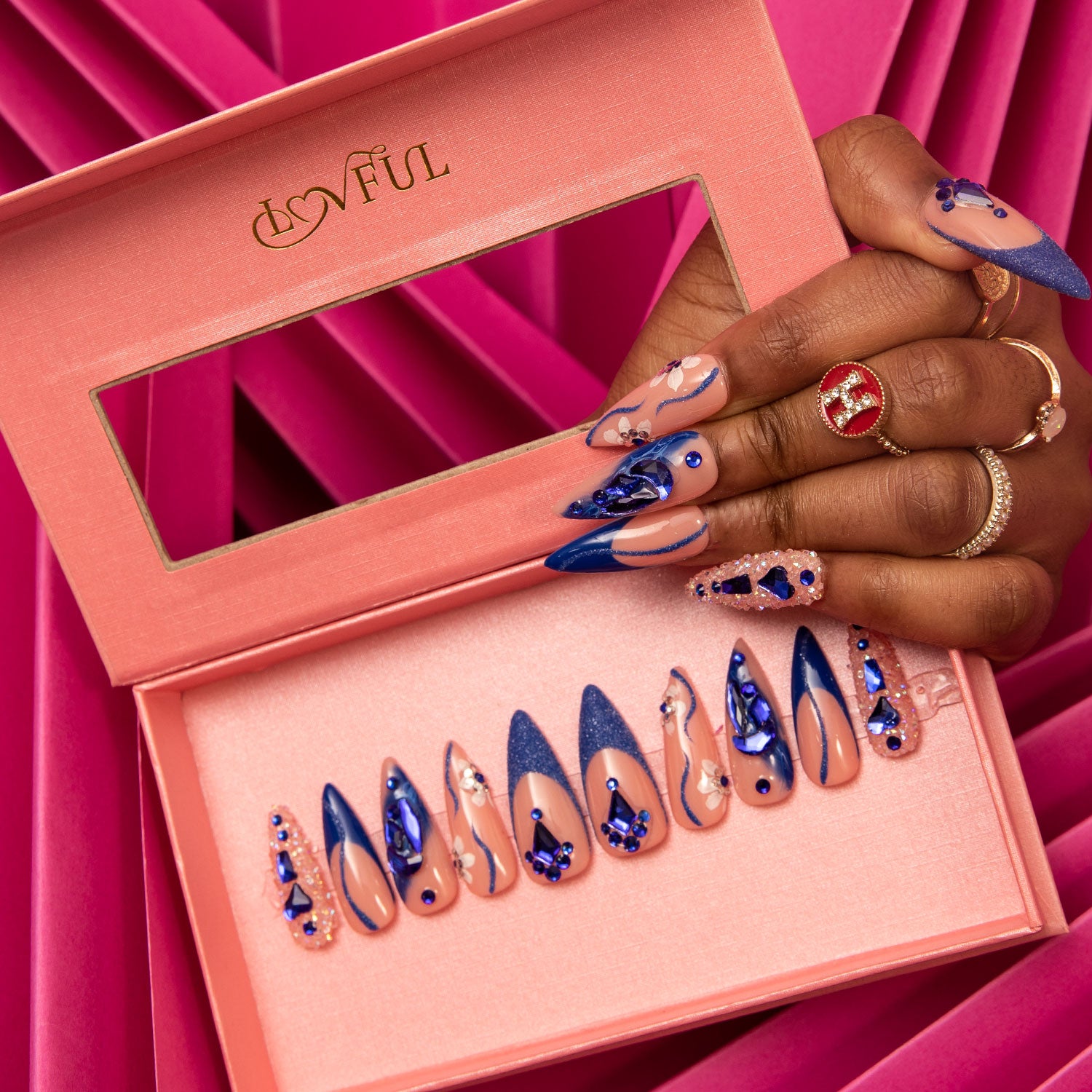 Open pink box displaying 'Blue Suede' press-on acrylic nails with blue French tips, curvy line design, crystal-like decorations, and blue heart-shaped gems. Hand with matching nails holds the box on a pink background.