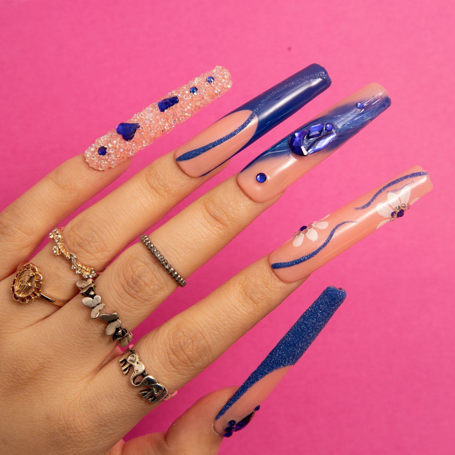 Hand with extended press-on nails featuring 'Blue Suede' design, showcasing blue French tips, crystal-like decorations, heart-shaped gems, and delicate curvy line designs against a pink background.