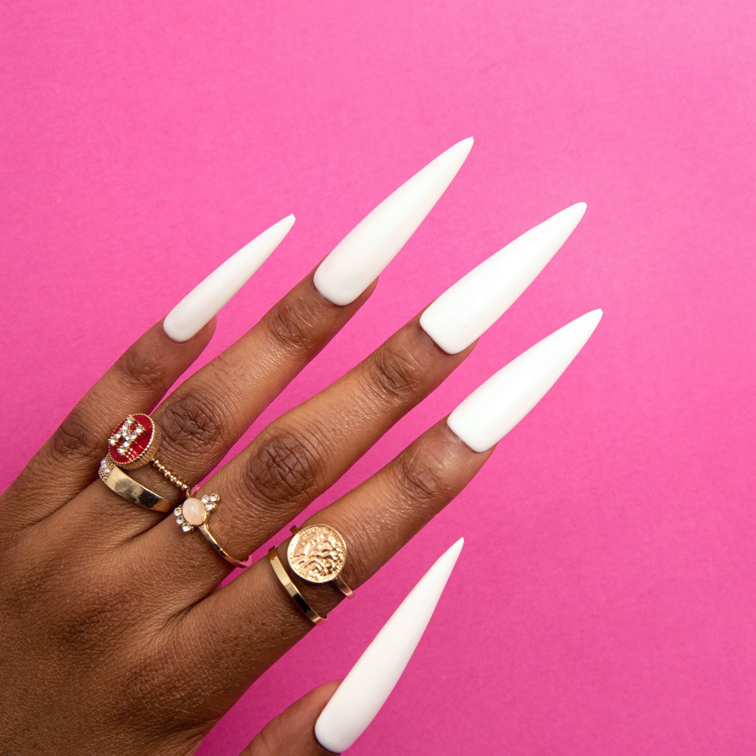 Hand with long, white stiletto-shaped press-on nails from Lovful.com, featuring multiple gold rings including floral and red gemstone designs, against a pink background.