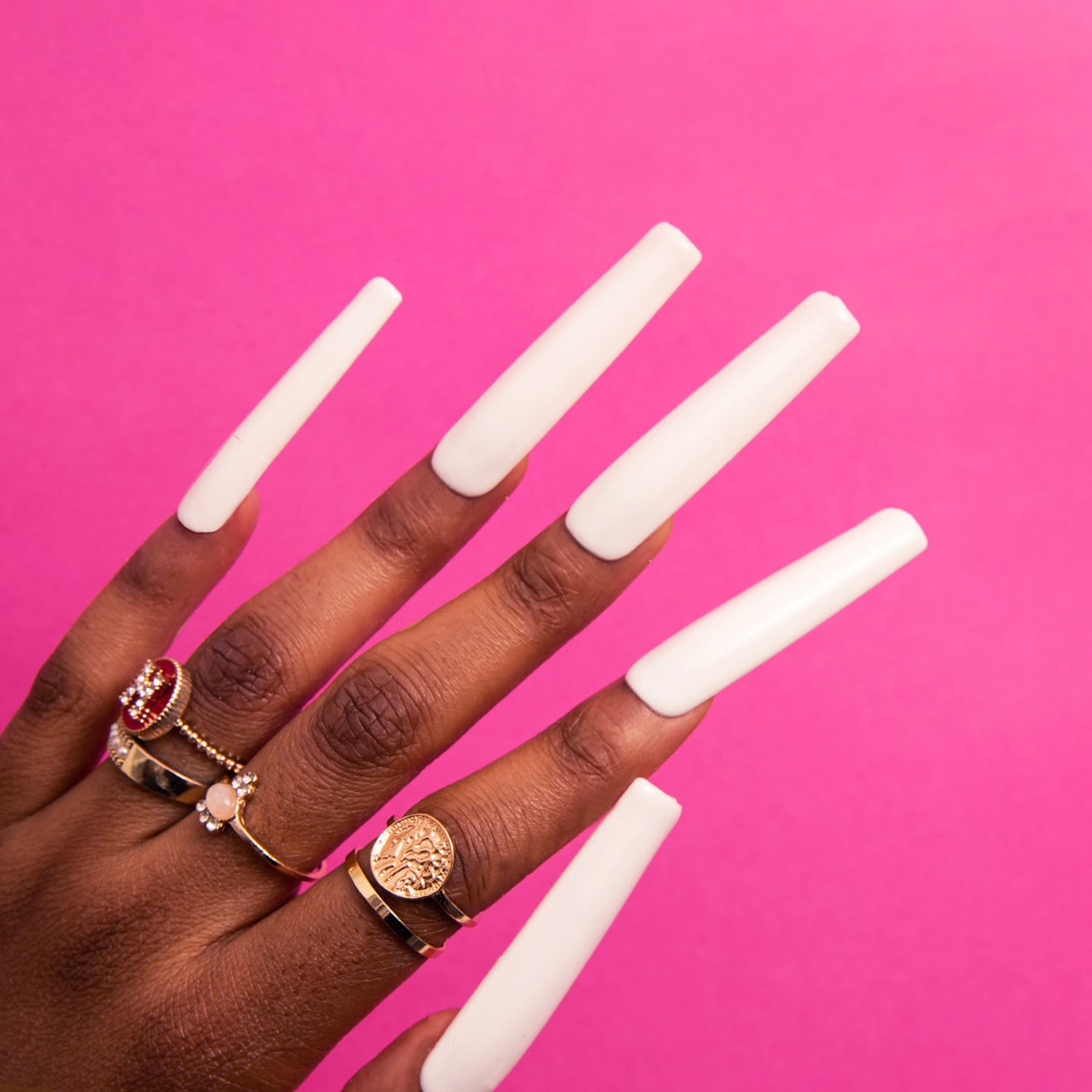 Hand with long, square-shaped, white press-on acrylic nails against a pink background, adorned with multiple rings including a red gemstone and gold rings. High-quality press-on nails by Lovful.com