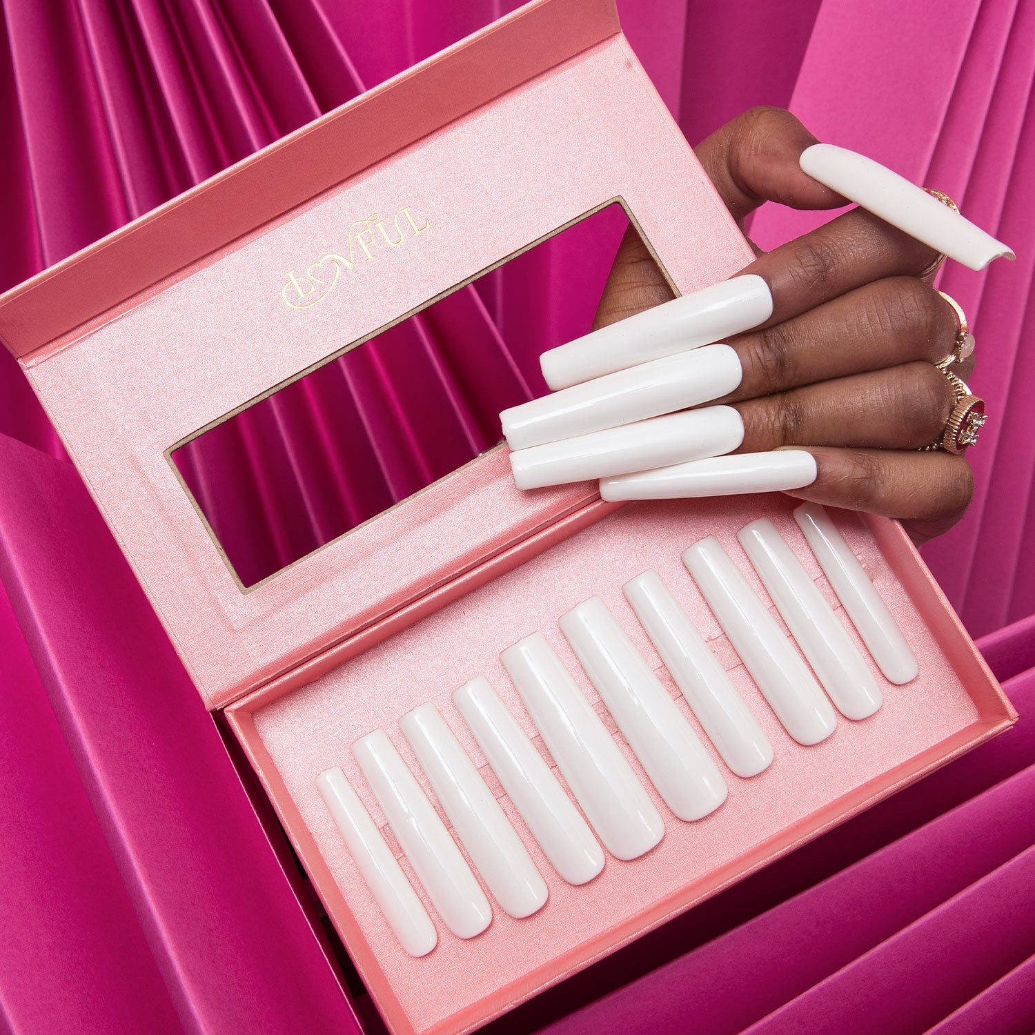 Hand displaying long white square-shaped press-on acrylic nails holding a Lovful pink box containing similar press-on nails against a pink pleated fabric background.