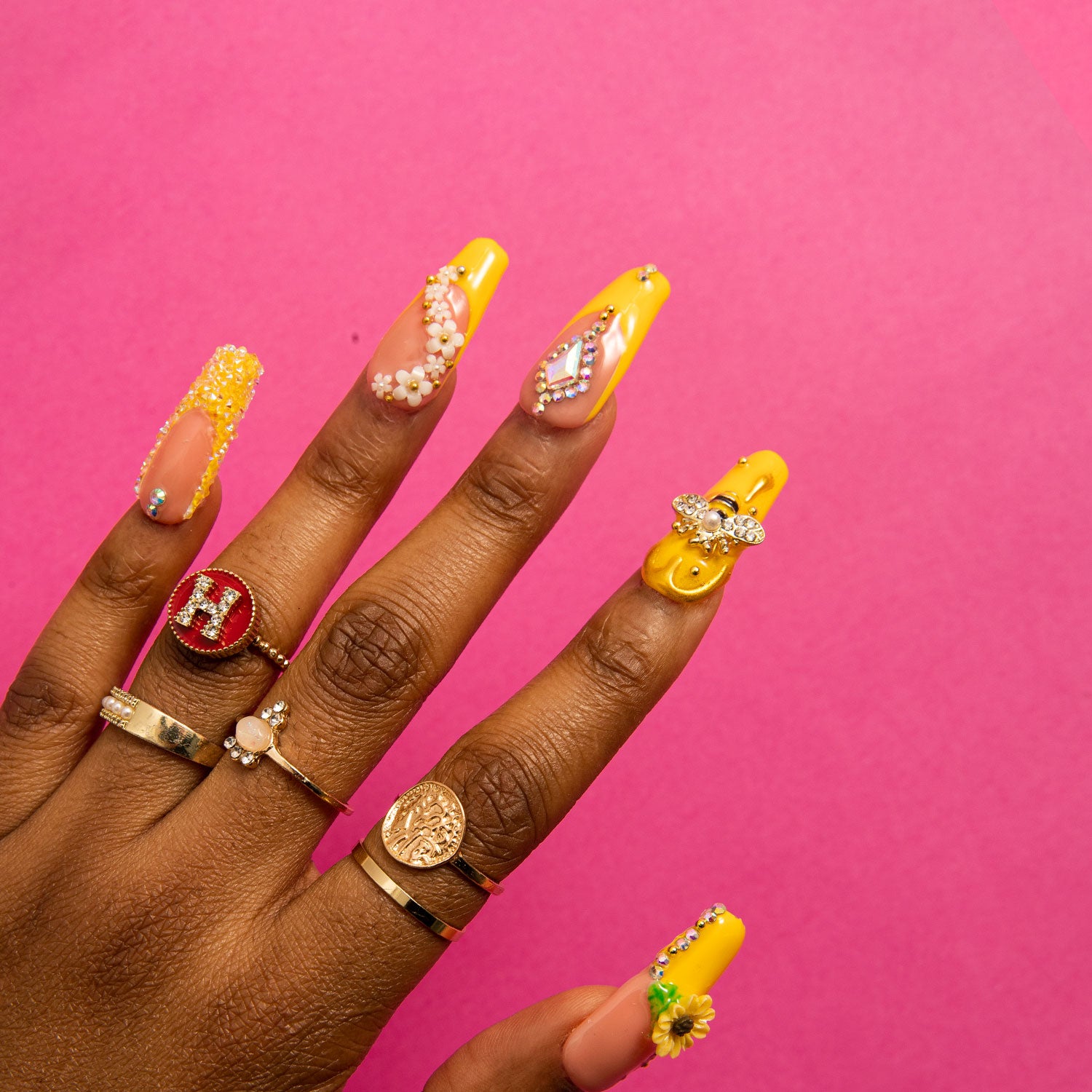 Hand with long coffin-shaped bright yellow press-on nails featuring sunflower patterns and bee accents, accessorized with multiple rings, against a pink background.