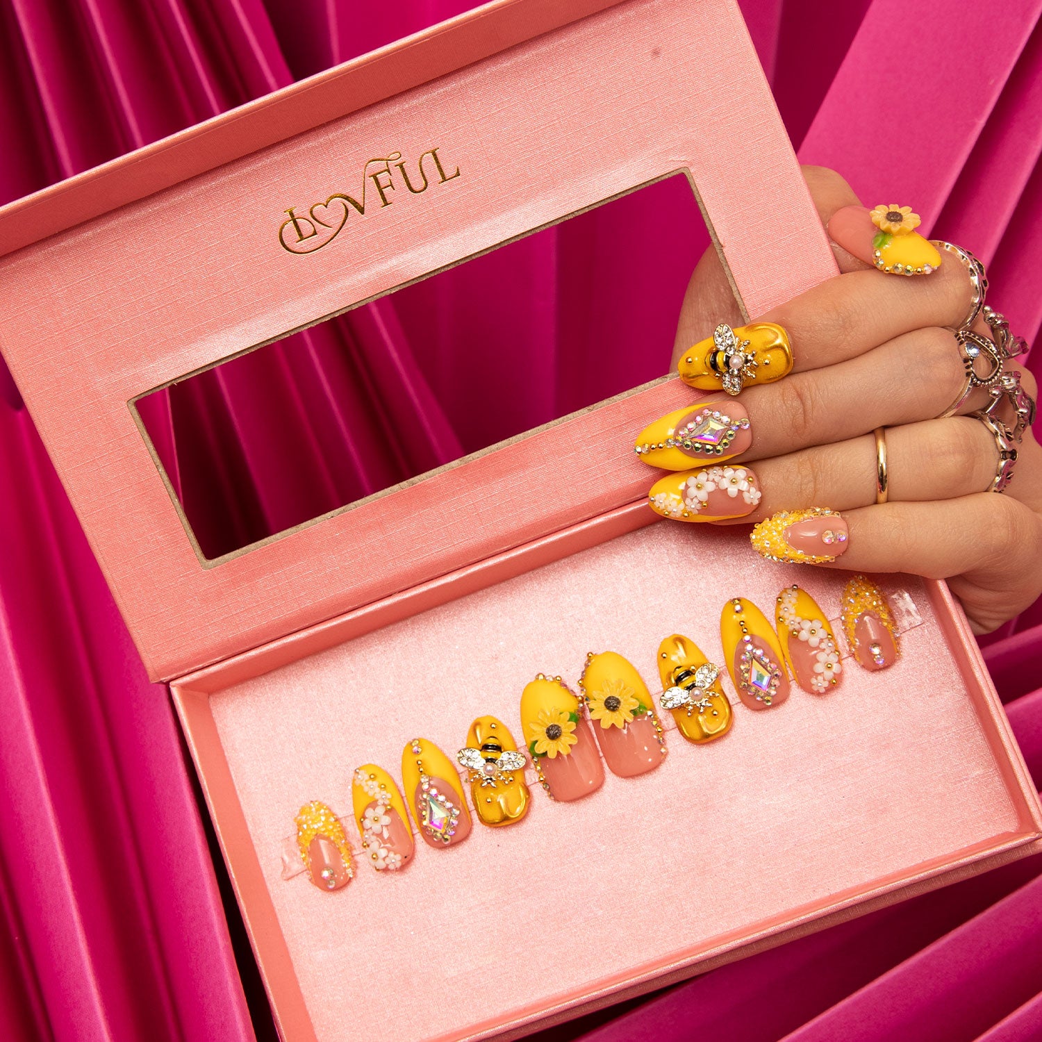 Lovful H101 Honey Bee Round press-on nails featuring a bright yellow design with charming sunflower patterns and adorable bee accents, displayed in a pink box with a clear window. A hand showcases the nails against a pink fabric background.