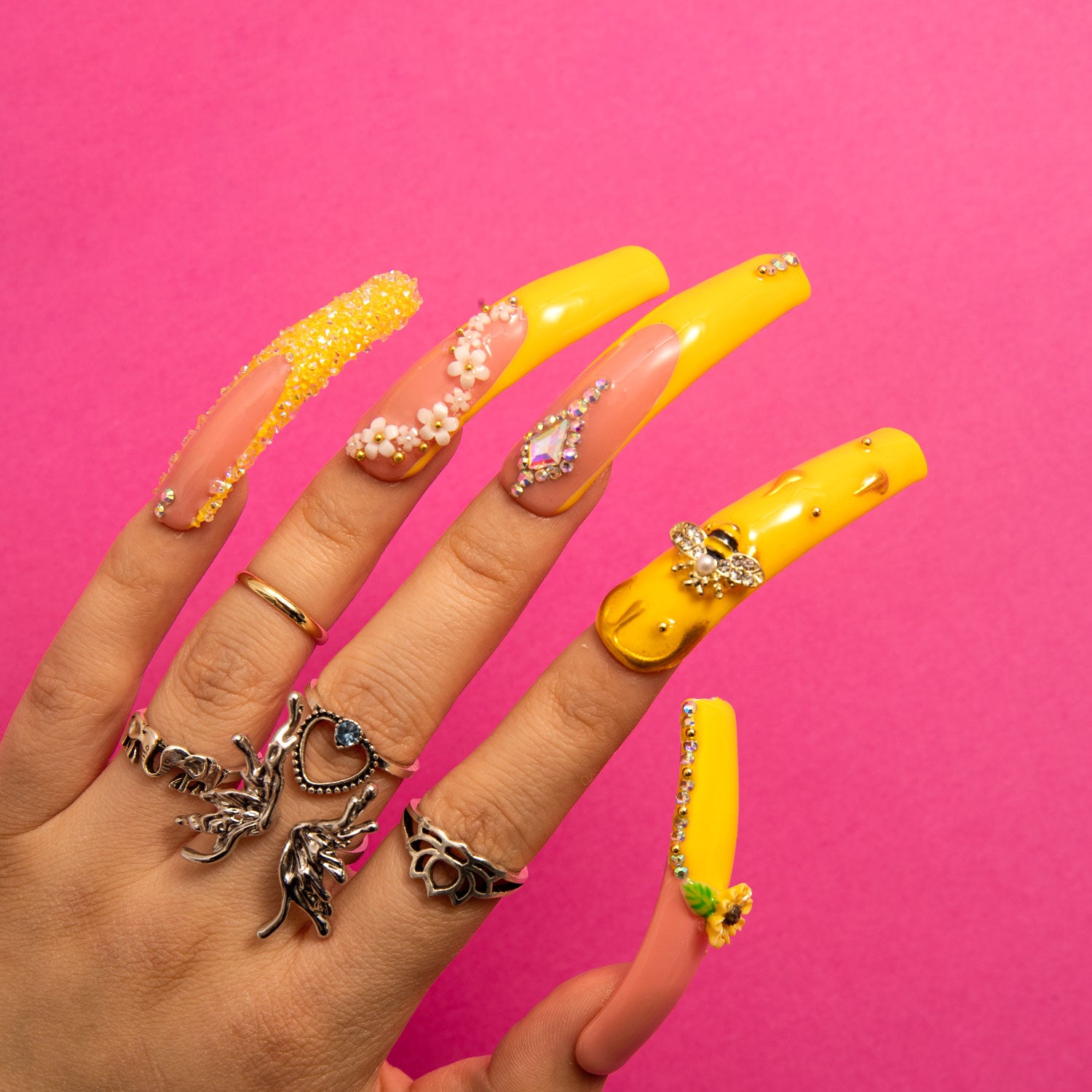 Hand with long, curved press-on acrylic nails in bright yellow and pink with sunflower patterns, bee accents, beads, flowers, and rhinestones, set against a pink background. Multiple rings with unique designs are also worn.