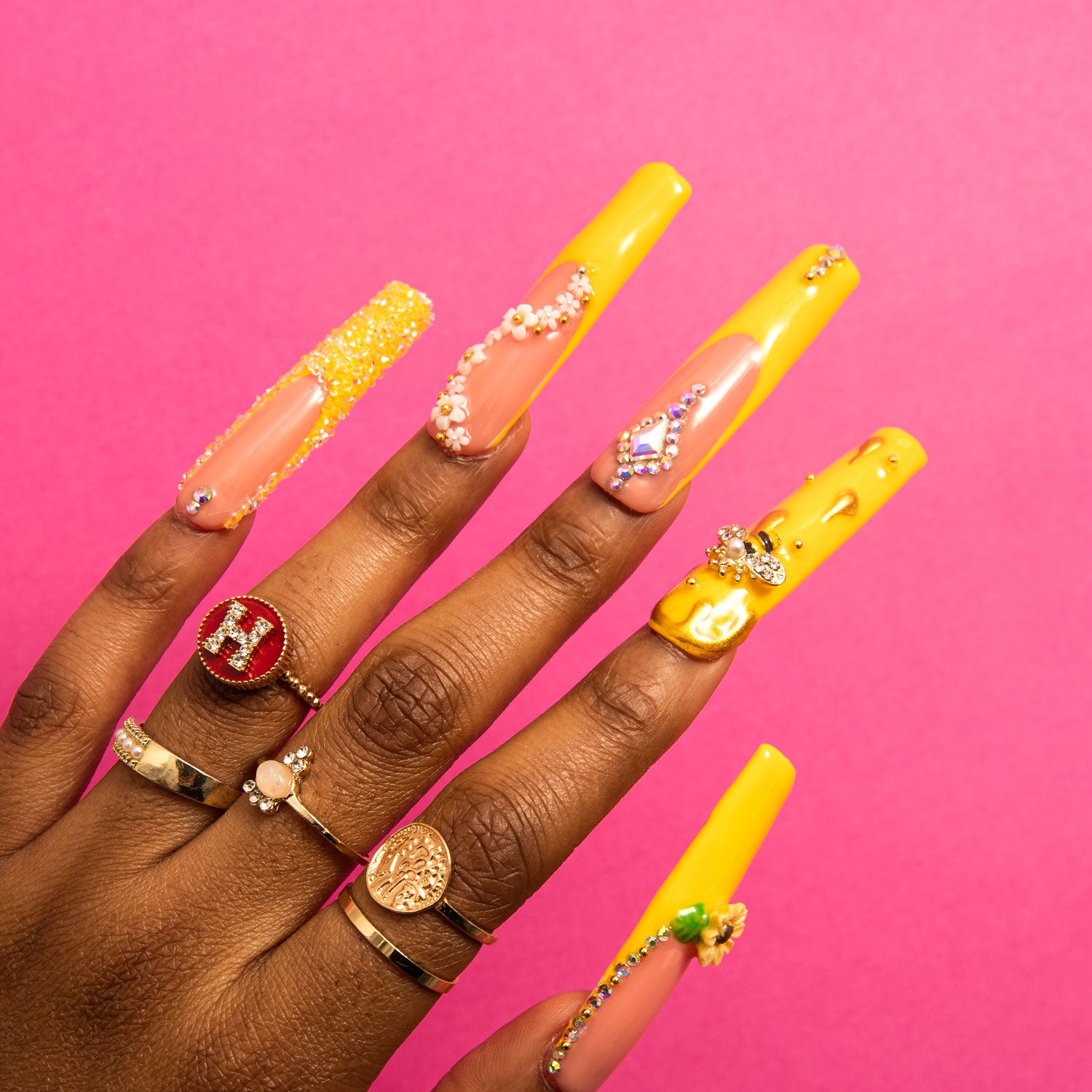 Hand with long square-shaped press-on acrylic nails in bright yellow with sunflower patterns, bee accents, and jeweled decorations against a pink background. Multiple rings on fingers, featuring designs like an 'H' and a flower.