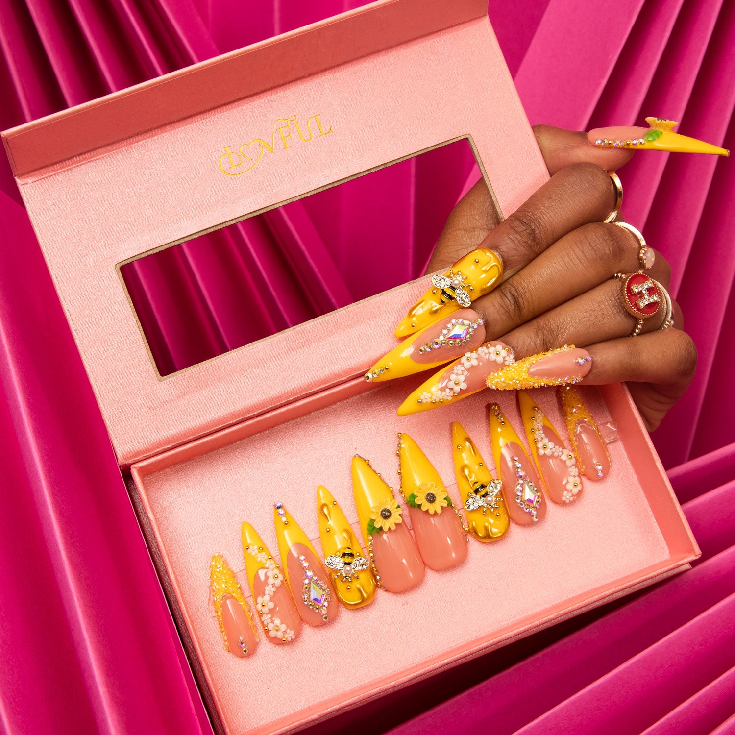 Bright yellow stiletto press-on nails with sunflower patterns and bee accents, displayed in a pink box. Hand with matching nails holding the box against a pink fabric background.