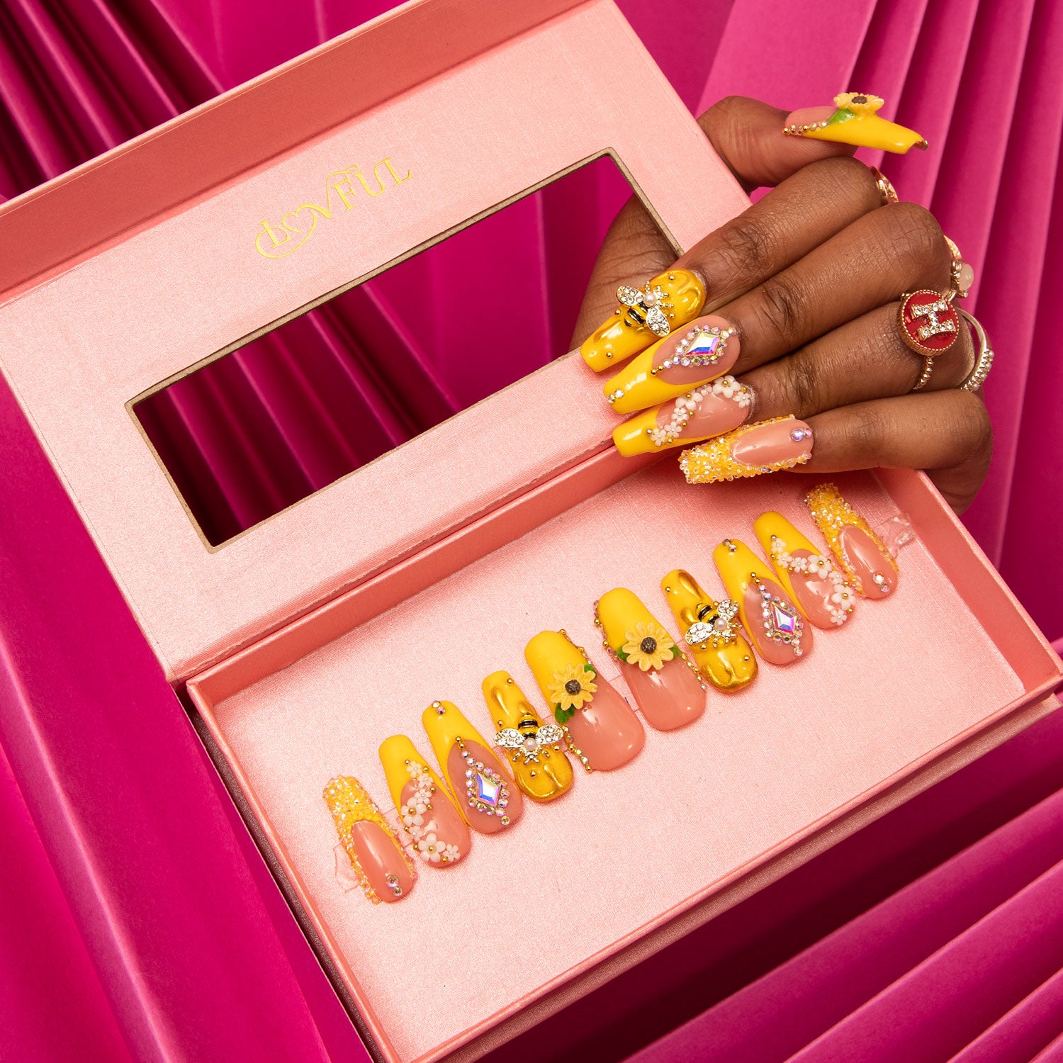 Bright yellow and pink press-on nails with sunflower patterns and bee accents by Lovful, displayed in a pink box. A hand with matching nail design is holding the box against a pink background.