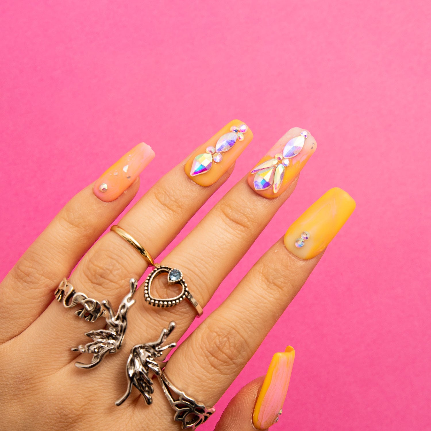 Acrylic nails painted in orange and yellow tones with rhinestone decorations, set against a pink background, and accessorized with silver rings including a heart-shaped ring.