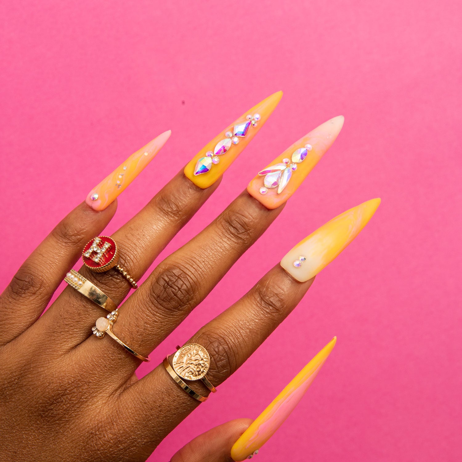 Hand with stiletto-shaped press-on nails in orange and yellow gradient with rhinestones and gold rings on a pink background, evoking the beauty of a sunset.