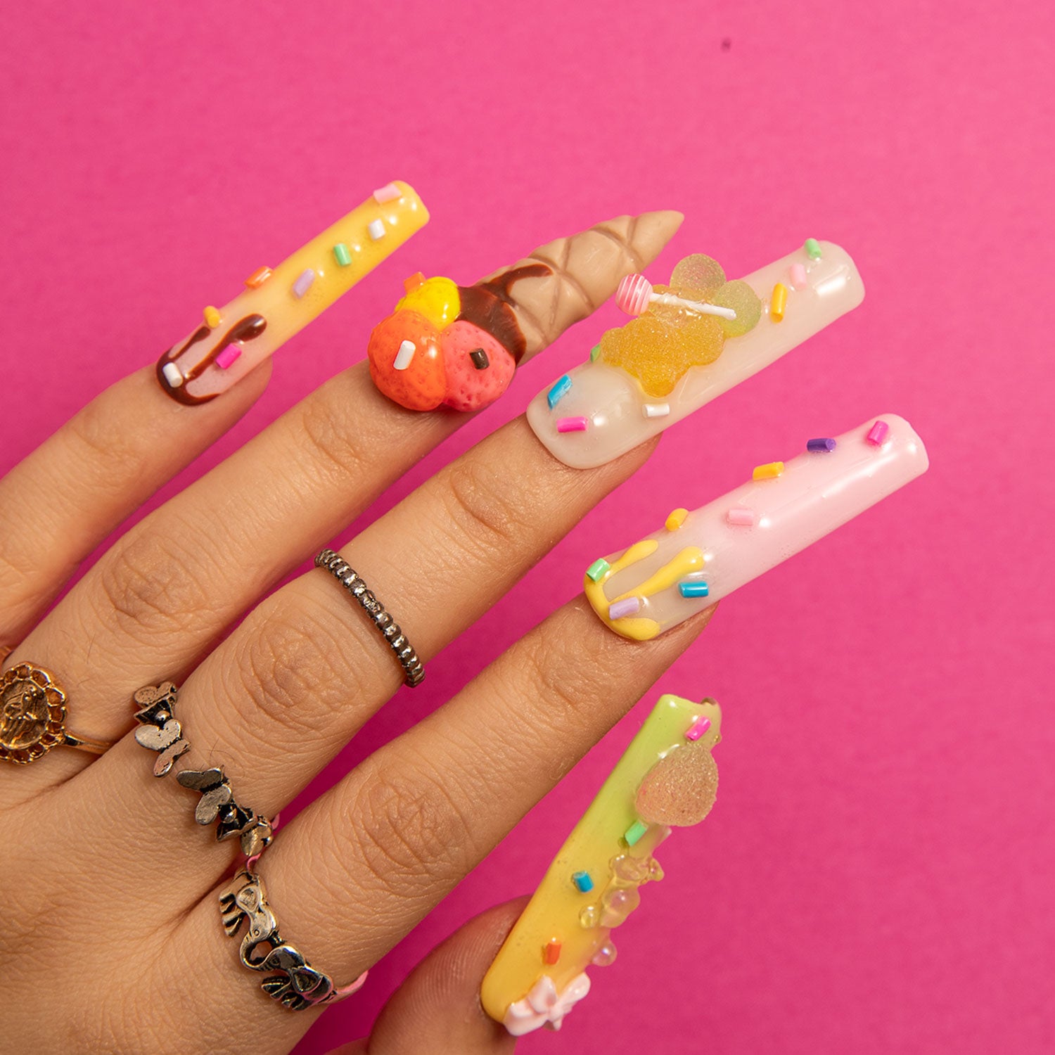 Square-shaped 3D decorated press-on nails with sprinkles, candy canes, and mini ice cream cones on a hand against a pink background