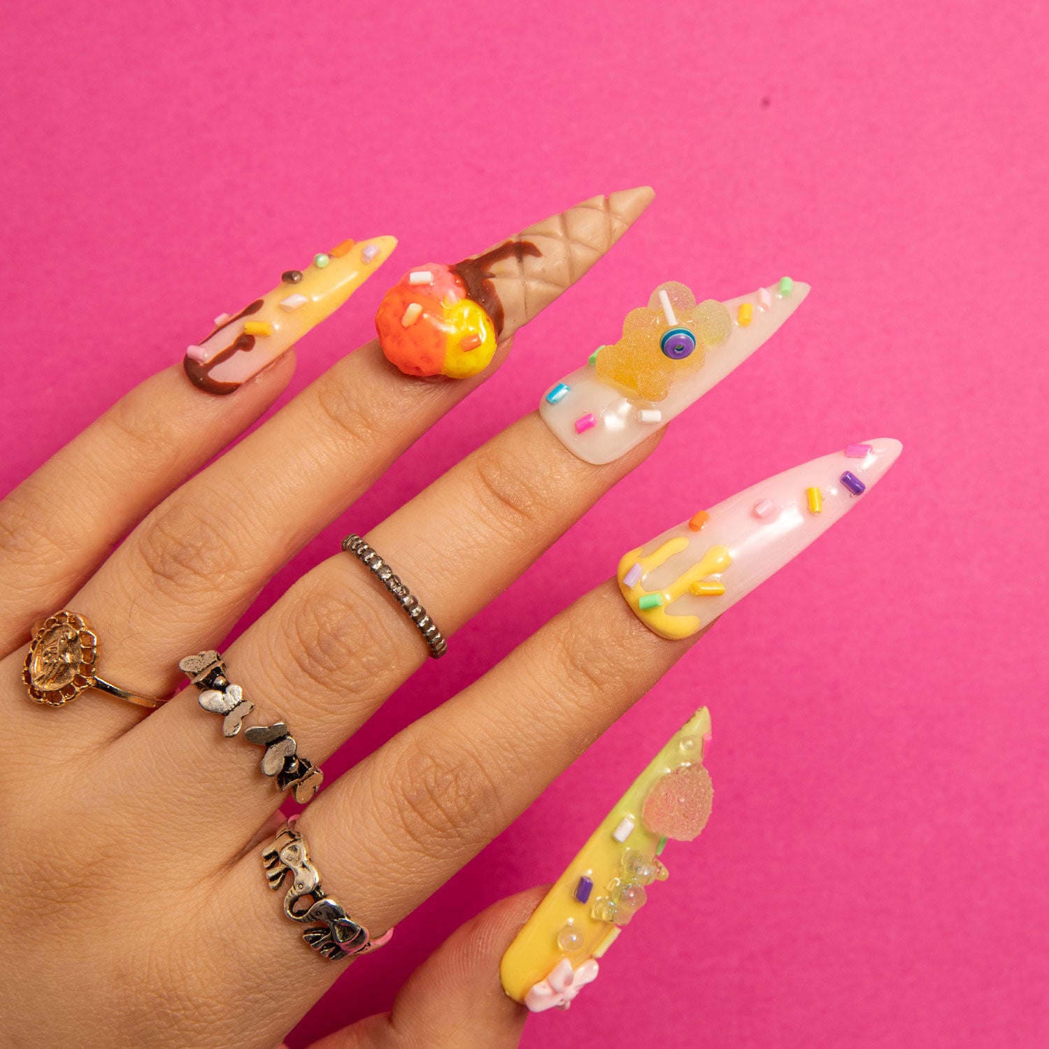 Hand with stiletto-shaped press-on nails featuring 3D decorations of sprinkles, candy canes, mini ice cream cones, and candy on a pink background