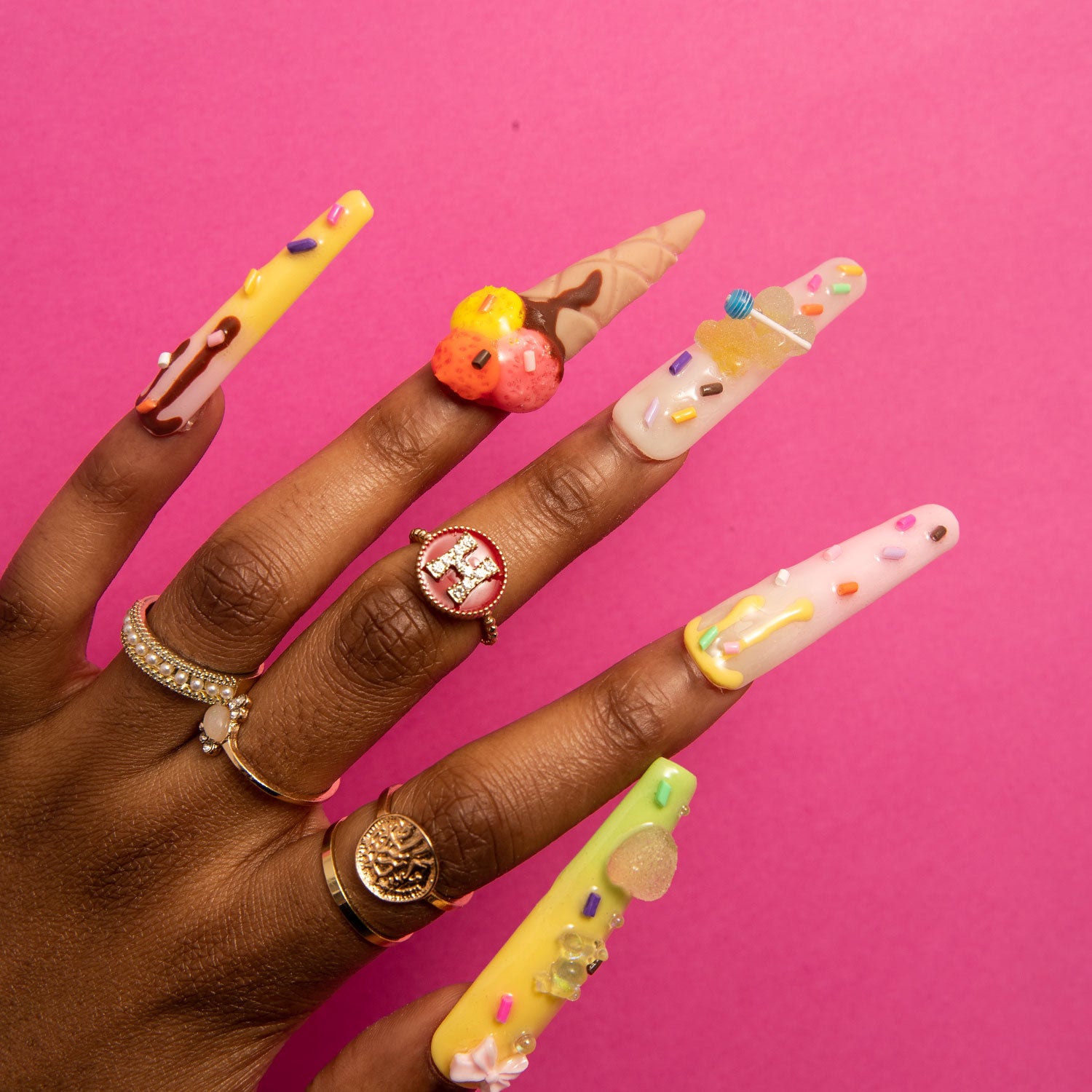 Hand with long coffin-shaped press-on nails featuring 3D decorations of sprinkles, candy canes, and mini ice cream cones against a pink background. Rings on fingers include a rose gold ring with a gemstone, a gold ring with a circular emblem, and a bee-shaped ring.