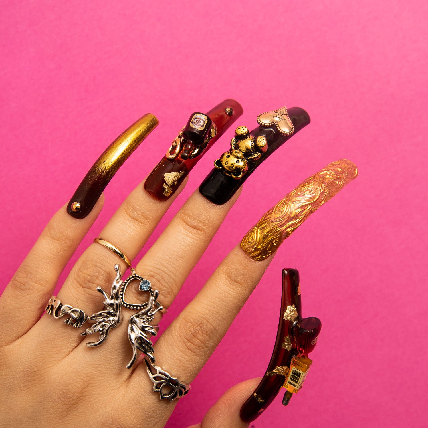 Hand showcasing 'Casino Party' curve press-on nails with black and gold color scheme, featuring bear and heart-shaped gems, and a wine bottle charm against a pink background. Silver rings with intricate designs complement the look.