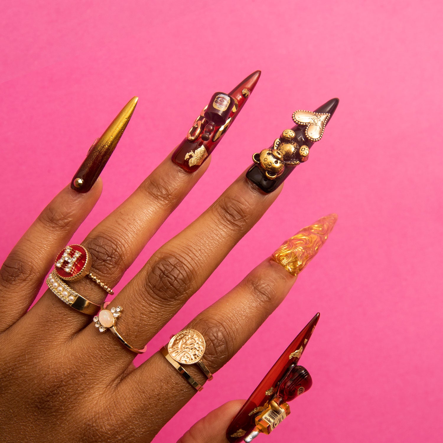 Hand wearing 'Casino Party' stiletto press-on nails with black and gold design featuring bear and heart-shaped gems, plus a wine bottle charm, accompanied by gold rings, against a pink background.