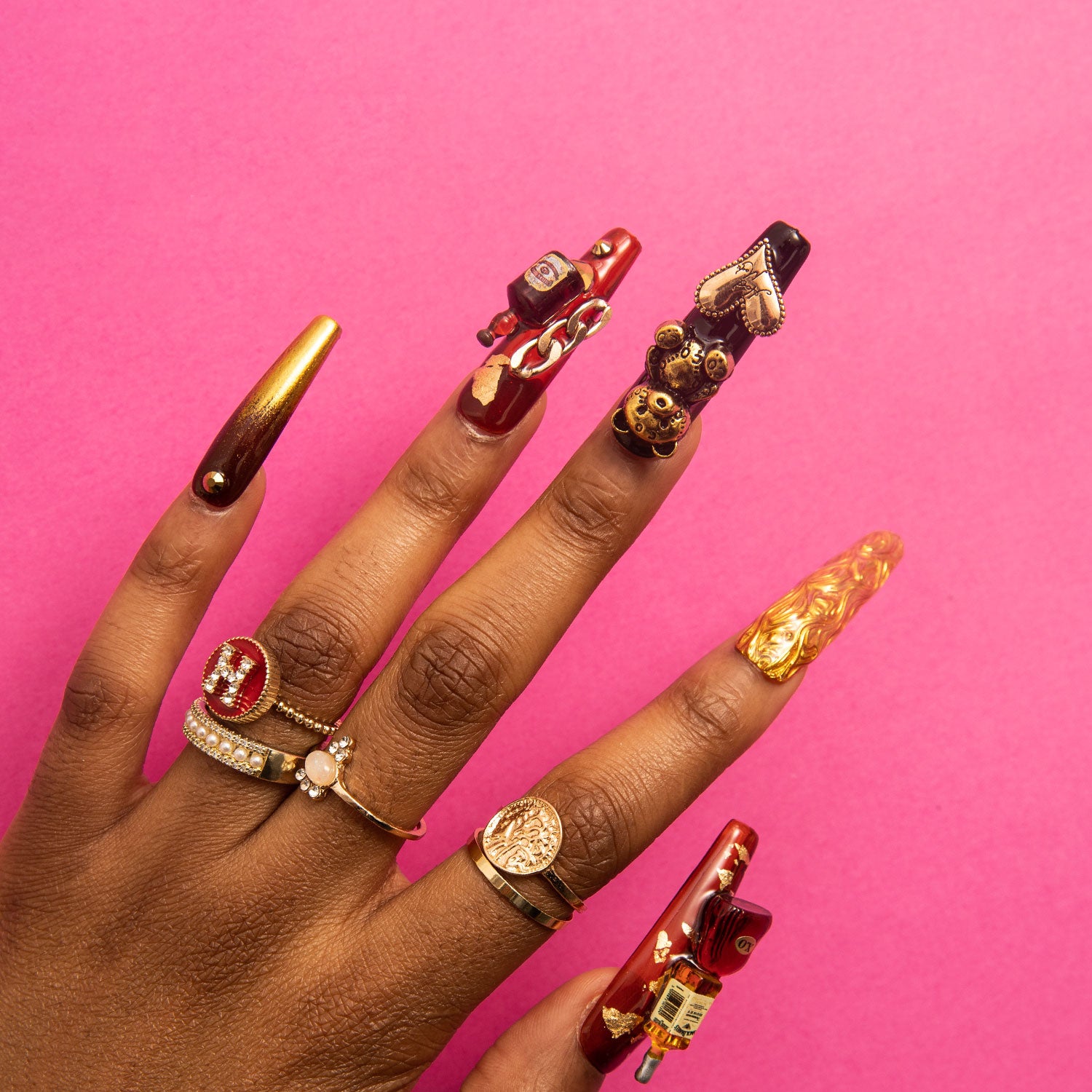 Hand showcasing luxurious Casino Party Press-On Nails with bear and heart-shaped gems, wine bottle charm, in black, gold, and red hues against a pink background