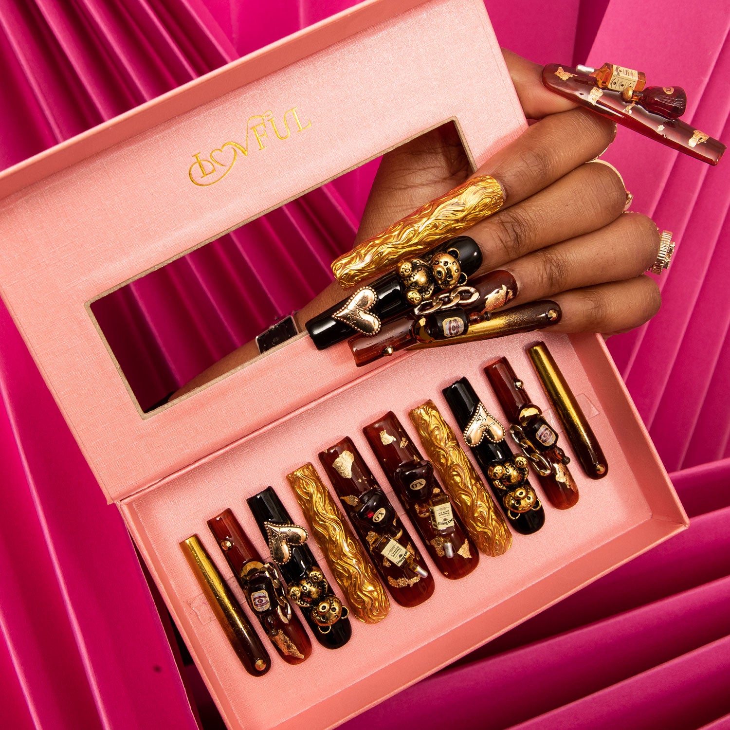 Hand showcasing 'Casino Party' press-on nails with black and gold designs, adorned with bear, heart-shaped gems, and a wine bottle charm, in a pink box with Lovful logo against a pink pleated fabric background.