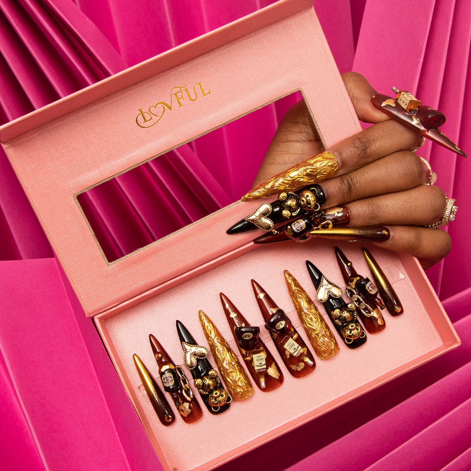 Set of eight stiletto-shaped press-on acrylic nails in luxurious black and gold with decorative gems and wine bottle charms in a pink box by Lovful, displayed on a vibrant pink background. A hand with matching nails holds the box.