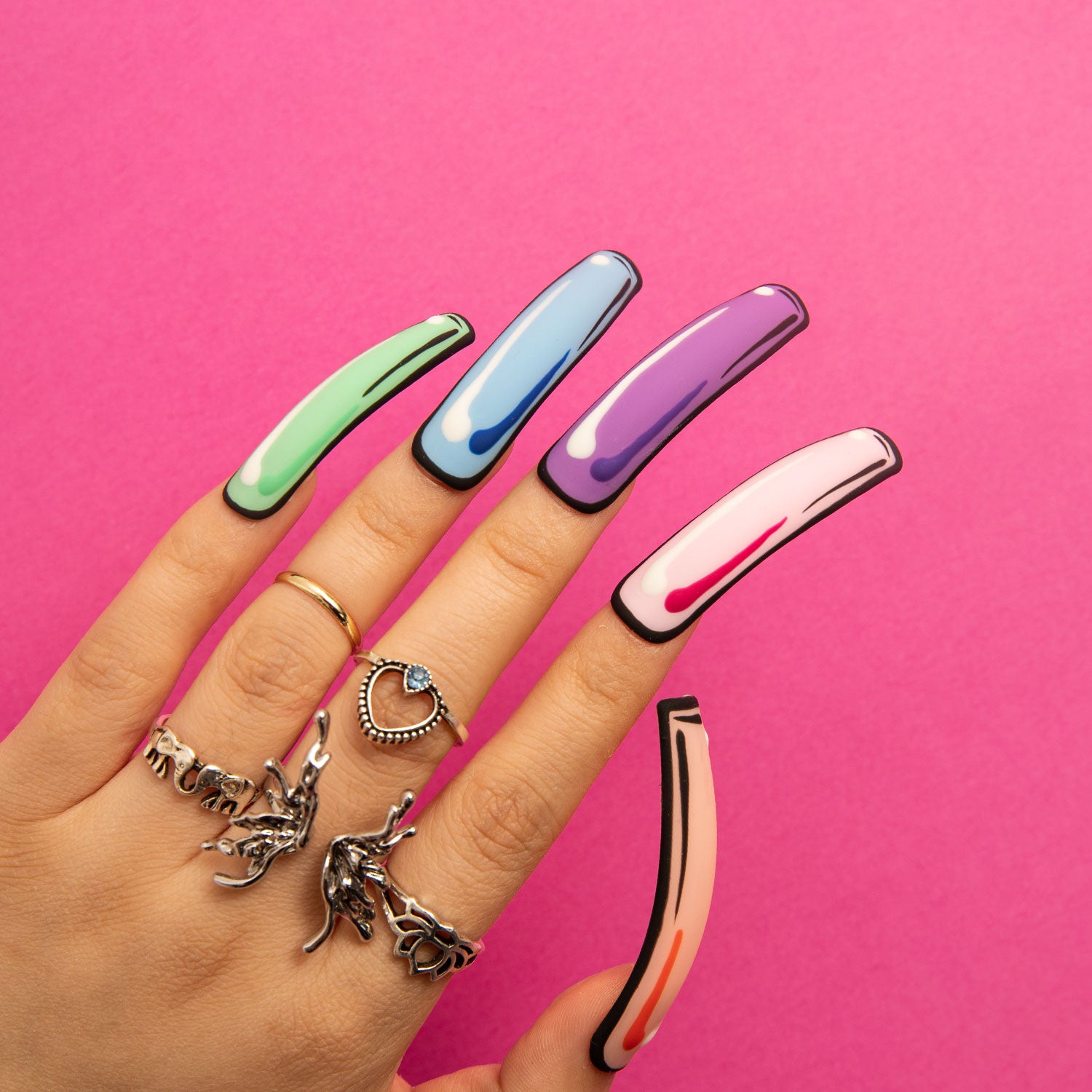 Hand wearing Lovful 'Colorful Pop' press-on nails with pop art design in green, blue, purple, white, and peach colors, set against a bright pink background. The hand features artistic rings.