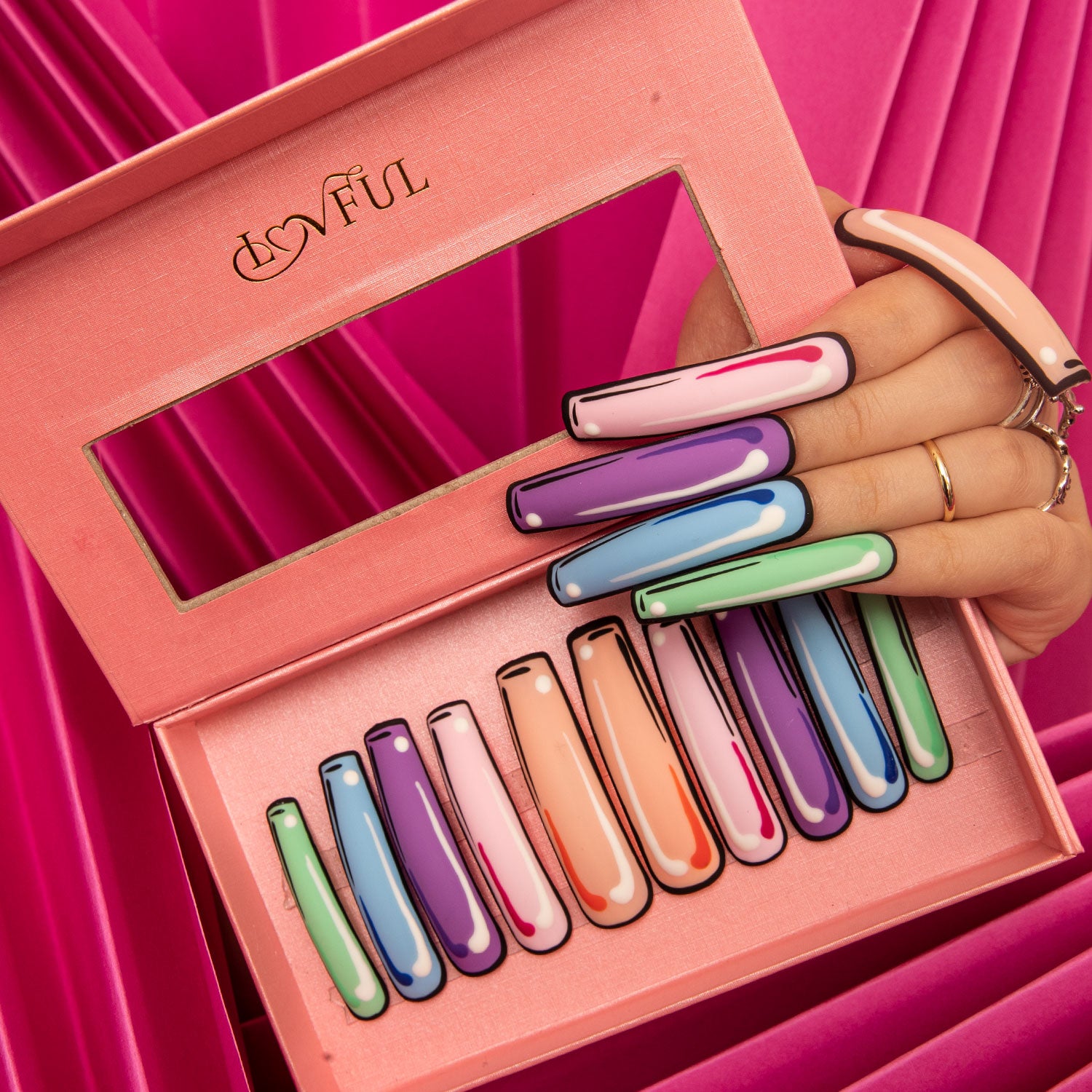 Colorful press-on acrylic nails set in a pink box from Lovful. The set features ten vibrant, pop art design nails in different colors, displayed against a pink background. A hand adorned with rings holds several nails.