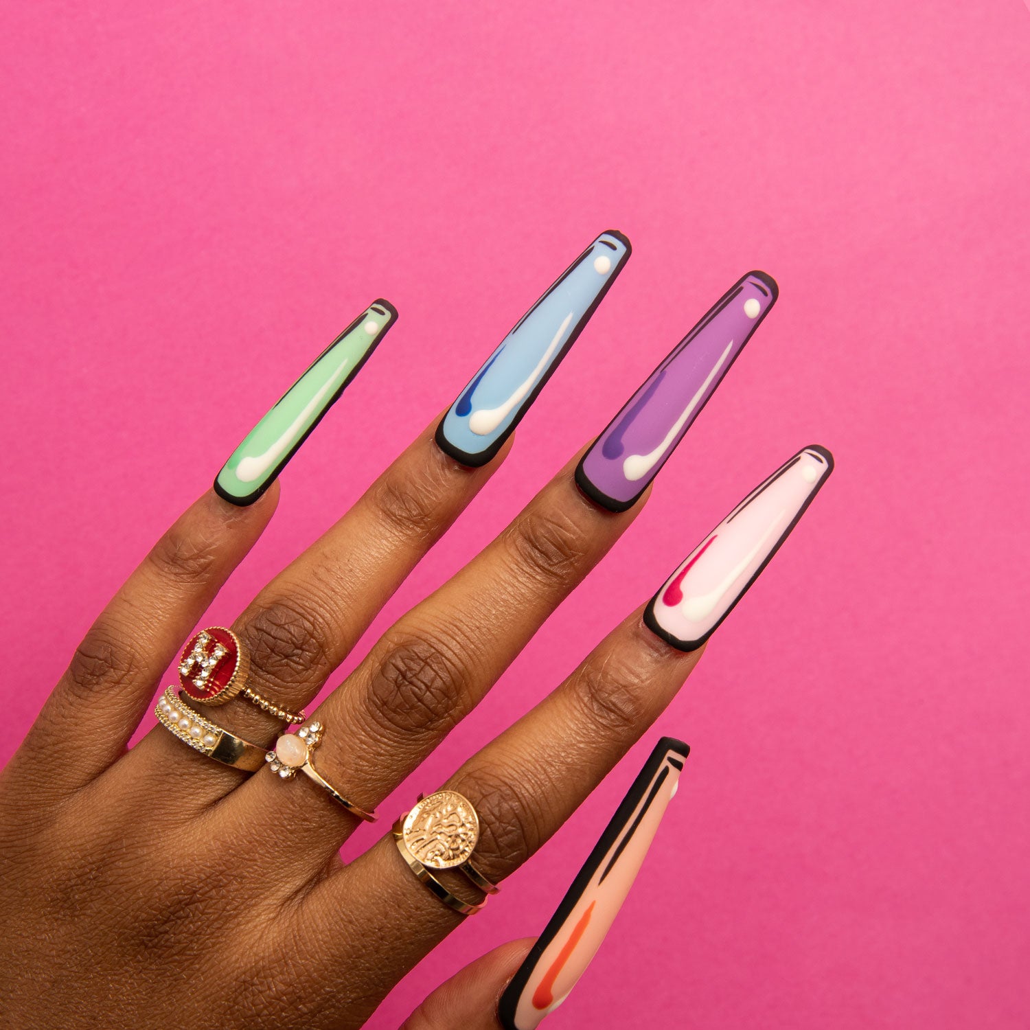 Hand with colorful coffin-shaped press-on nails featuring pop art design, including green, blue, purple, pink, and orange colors, outlined in black, against a pink background. Adorned with gold and gemstone rings. Lovful.com.