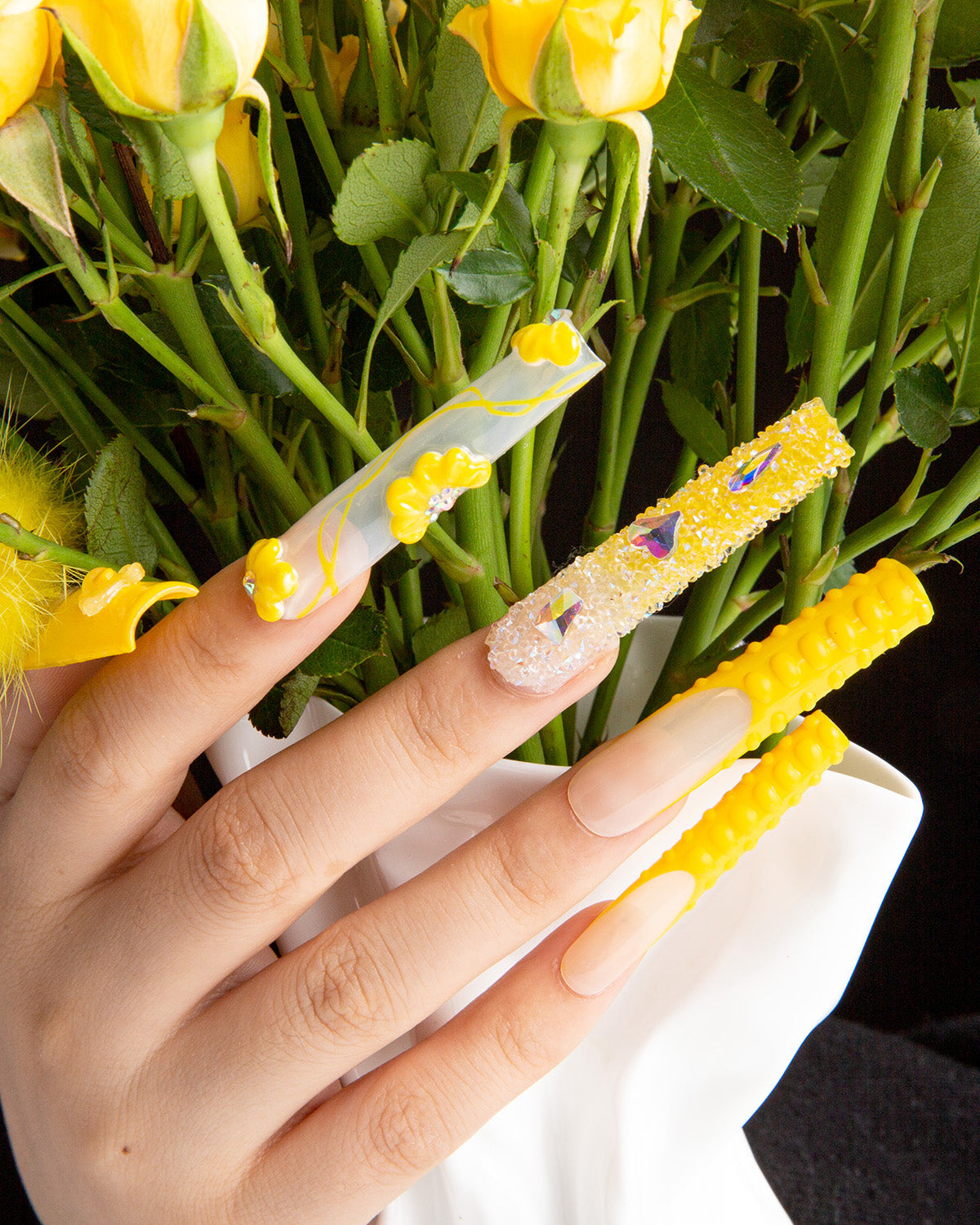 Hand showcasing 24 sunny yellow press-on acrylic nails from Lovful's 'Yummy' collection, featuring unique designs like marbling, decorative elements, and detachable fluffy balls, accentuated by yellow flowers in the background.