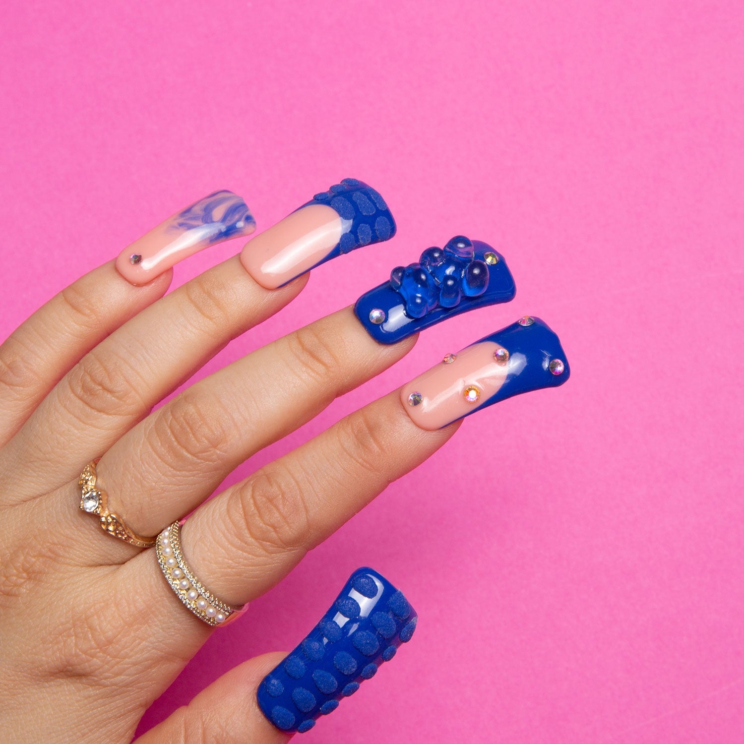 A hand with Little Blue Bear Rhinestone French tip nails, featuring blue hue, 3D gummy bears, shiny rhinestones, and hand-drawn 3D dots against a pink background.