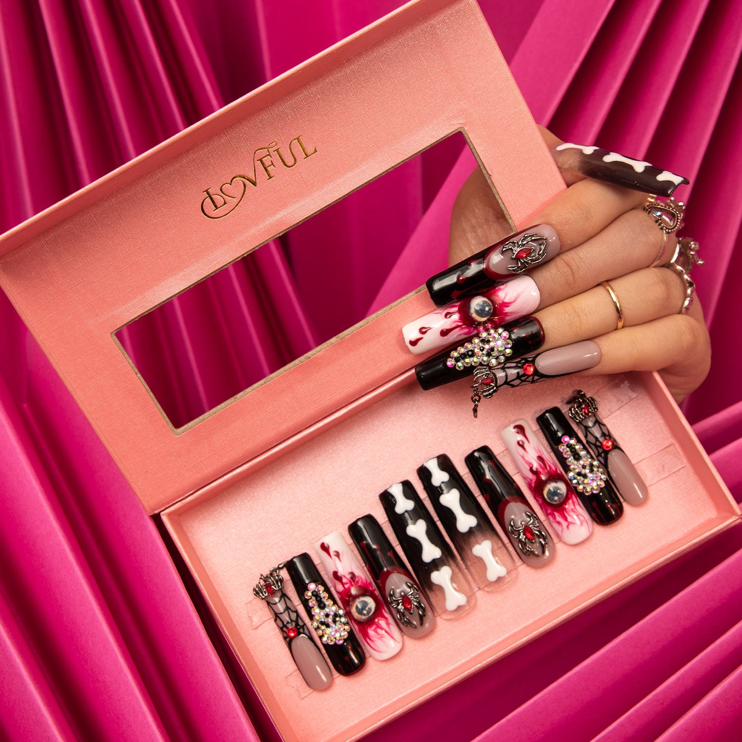 Hand with Halloween-themed press-on nails including eyes, ghosts, bones, and blood patterns holding a pink box containing similar nail designs from Lovful against a pink fabric background.