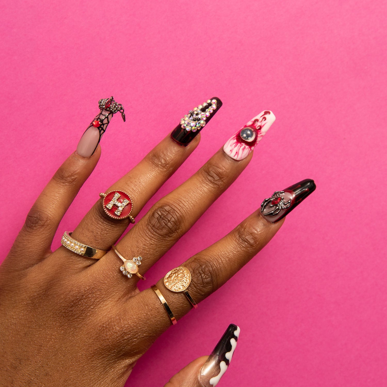 Hand with long coffin-shaped press-on nails with Halloween-themed designs, featuring ghost faces, skeletal bones, and sinister eyes in black and blood-red, against a pink background. The hand is accessorized with gold rings.