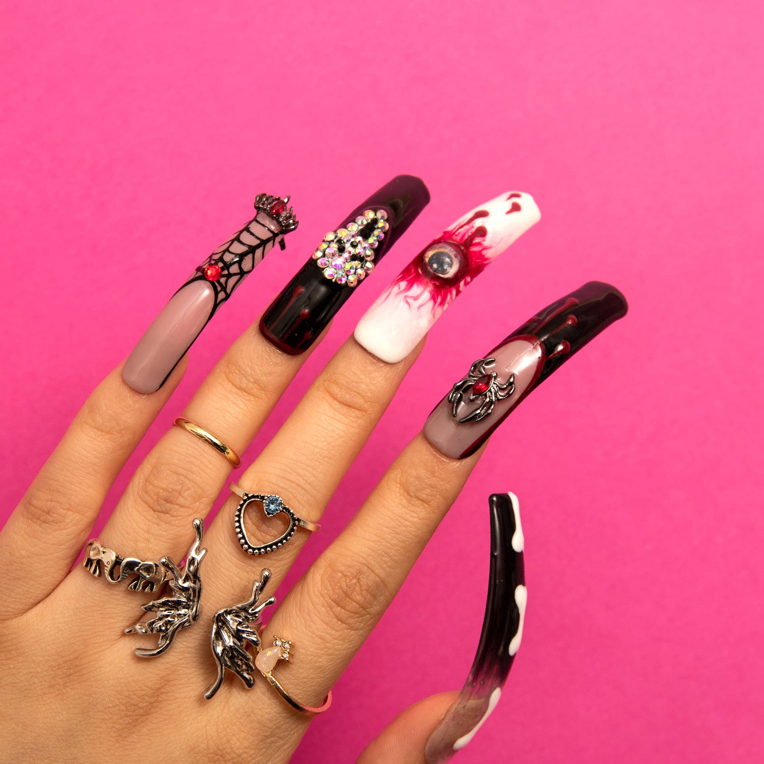 Hand with Halloween-themed press-on acrylic nails featuring designs of a spider web, bejeweled ornament, bloody eyeball, and sinister skull from the 'Final Destination' collection, against a pink background and adorned with various rings.