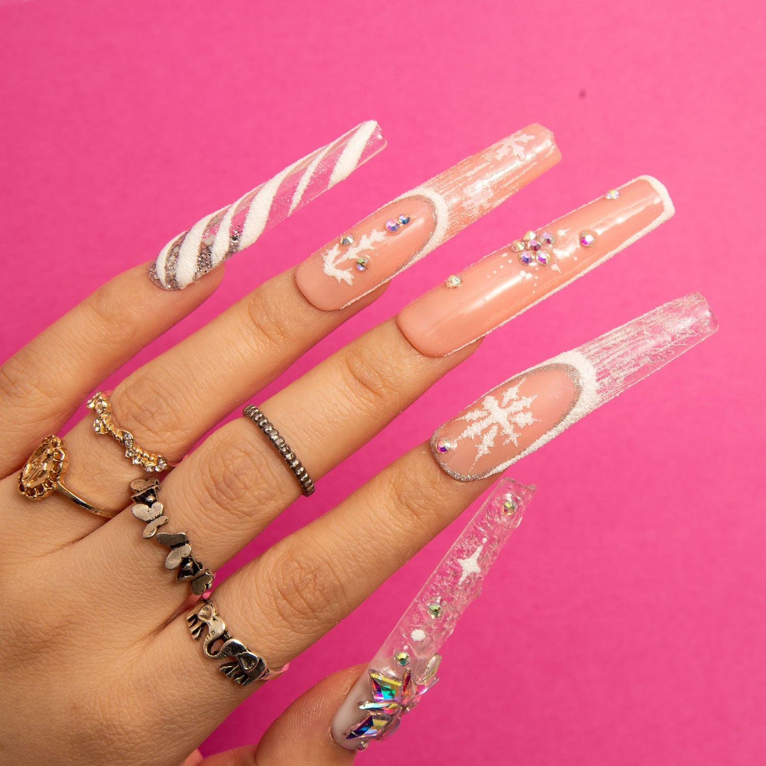 Hand with five press-on acrylic nails featuring winter-themed designs with white snowflakes, glitter, and rhinestones against a pink background