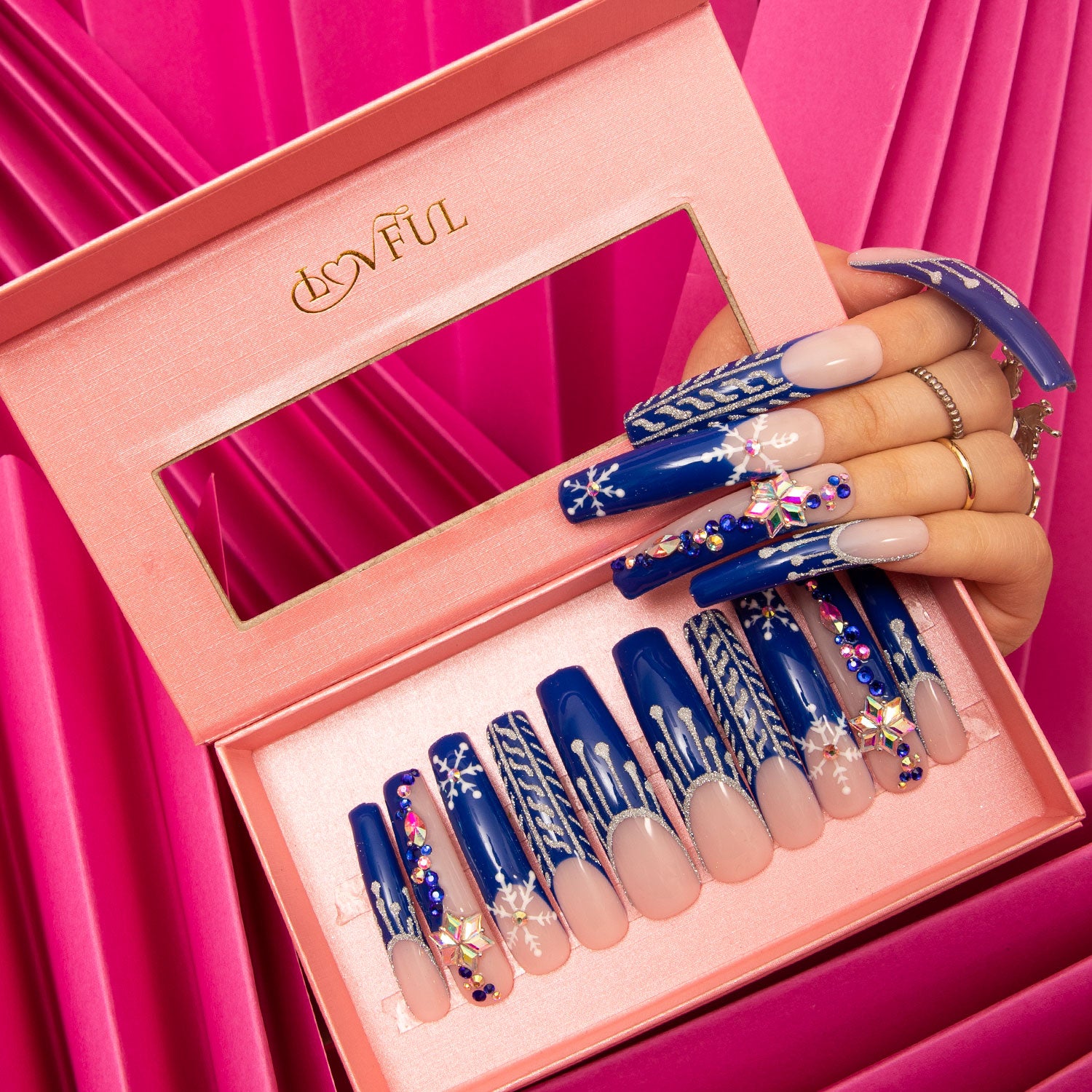 Lovful Snow Waltz press-on nails with blue jewelry-colored French tips and white snowflakes in a pink box. Hand showcasing the nails against a pink fabric background.