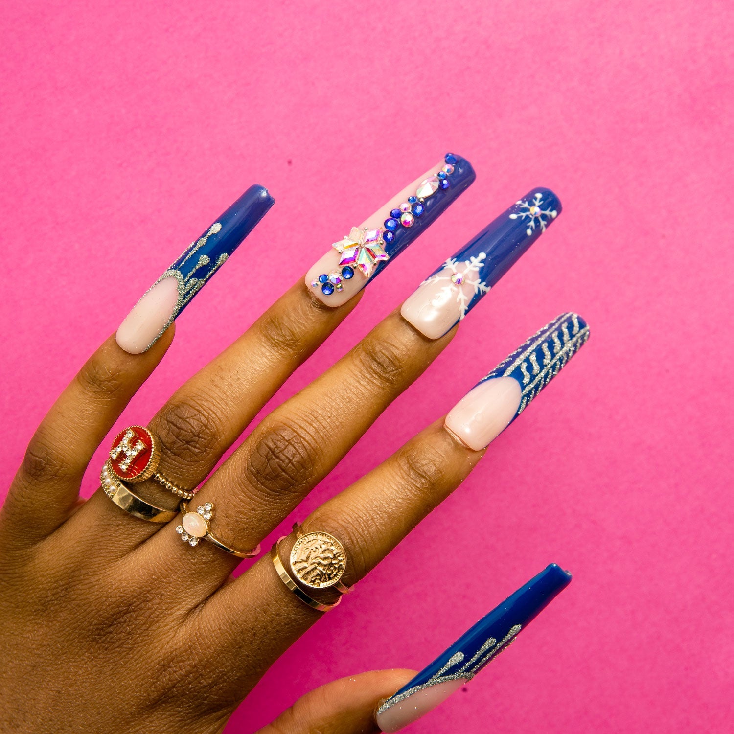 Hand with long square-shaped blue French tip press-on nails featuring winter-themed designs, including white snowflakes and jewelry-colored accents, against a pink background. The person is also wearing gold rings.