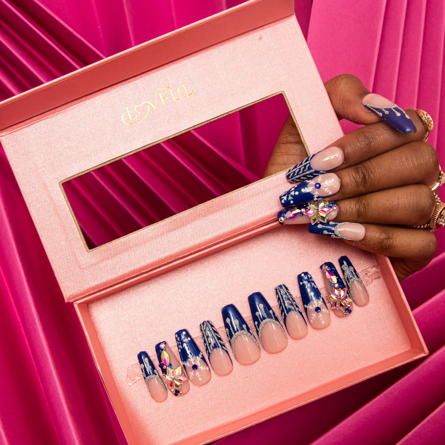 Snow Waltz Press-On Nails with blue jewelry-colored French tips and white snowflakes displayed in an elegant pink box. Hand showcasing the decorated nails alongside the box against a pink fabric background.