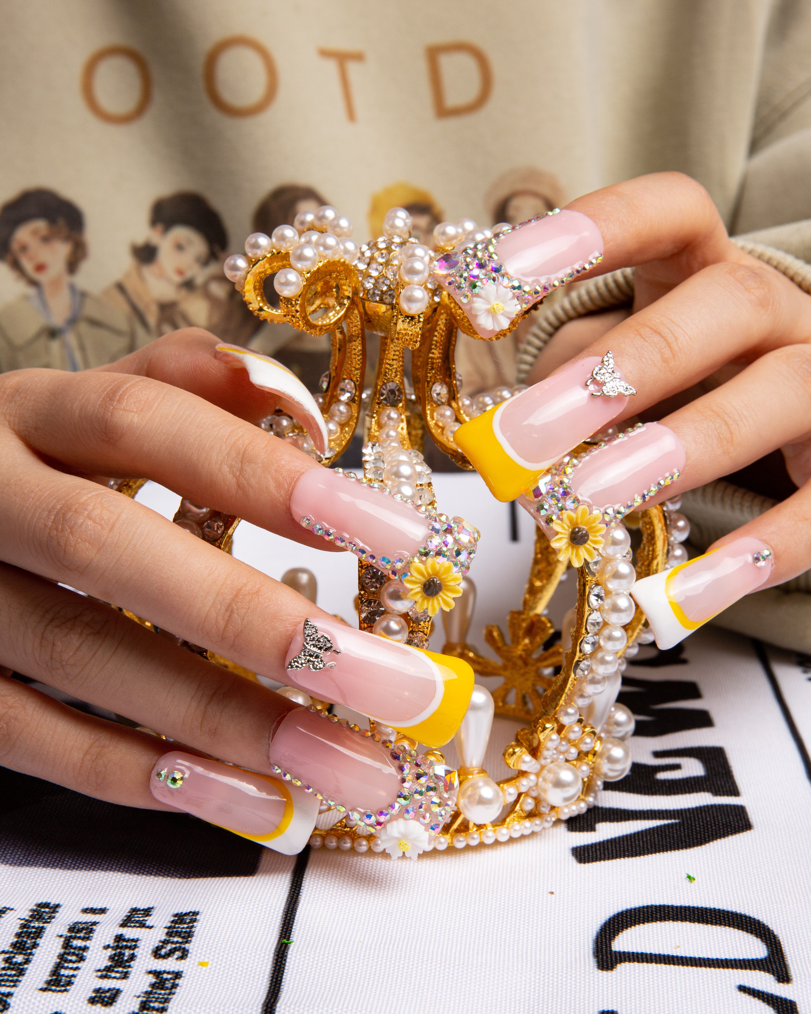 Hands displaying Sunflower press-on nails with yellow French tips, sunflower accents, and rhinestones, holding an ornate golden crown with pearls. Lovful's high-quality acrylic nails.