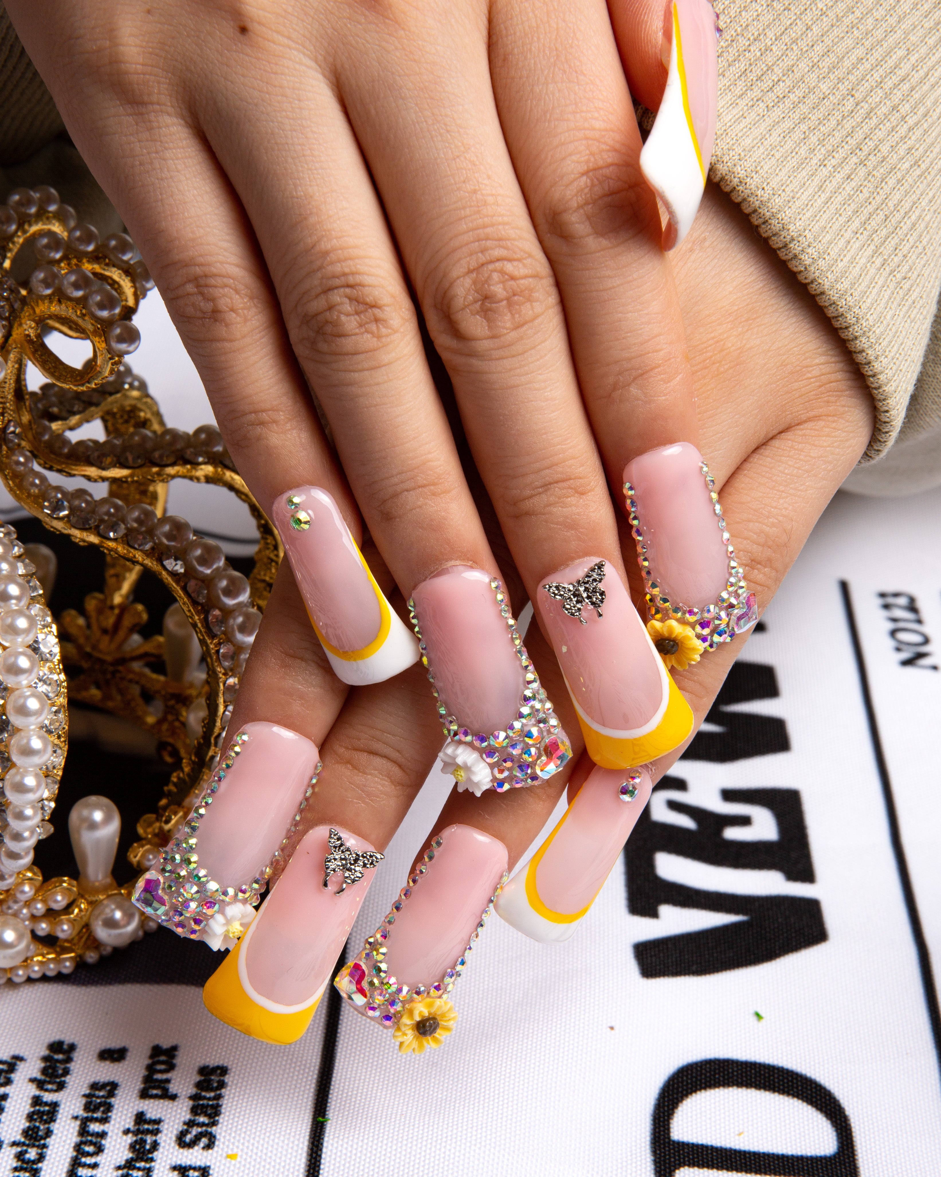 Press-on nails with a sunflower design in vibrant yellow and white French tips, decorated with rhinestones and butterfly decals, against an elegant background with pearls and a golden ornament.