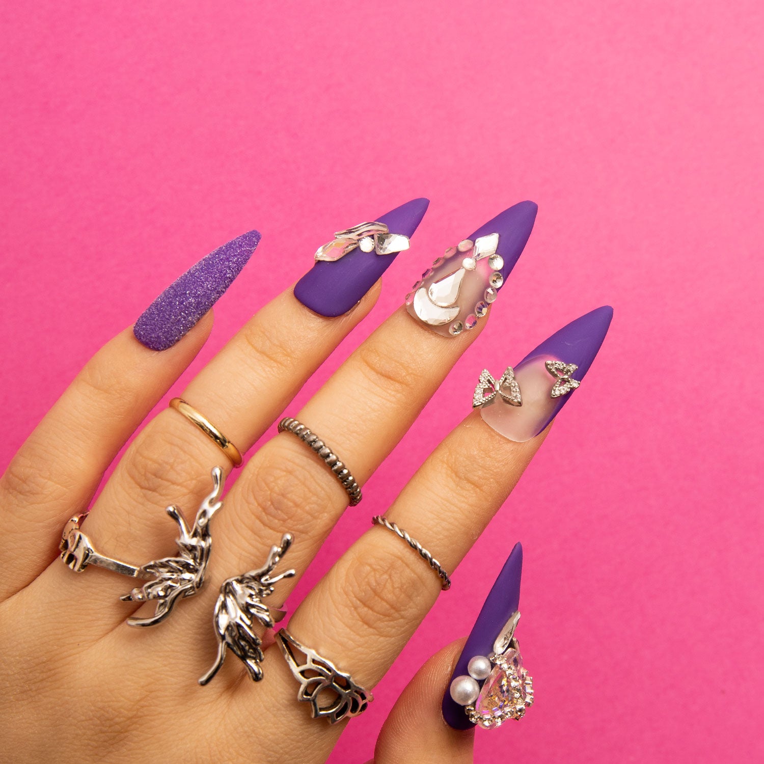 Mystique Veil Stiletto press-on nails with deep purple base, butterfly decorations, gem accents, and glitter overlay on a hand wearing silver rings, pink background.